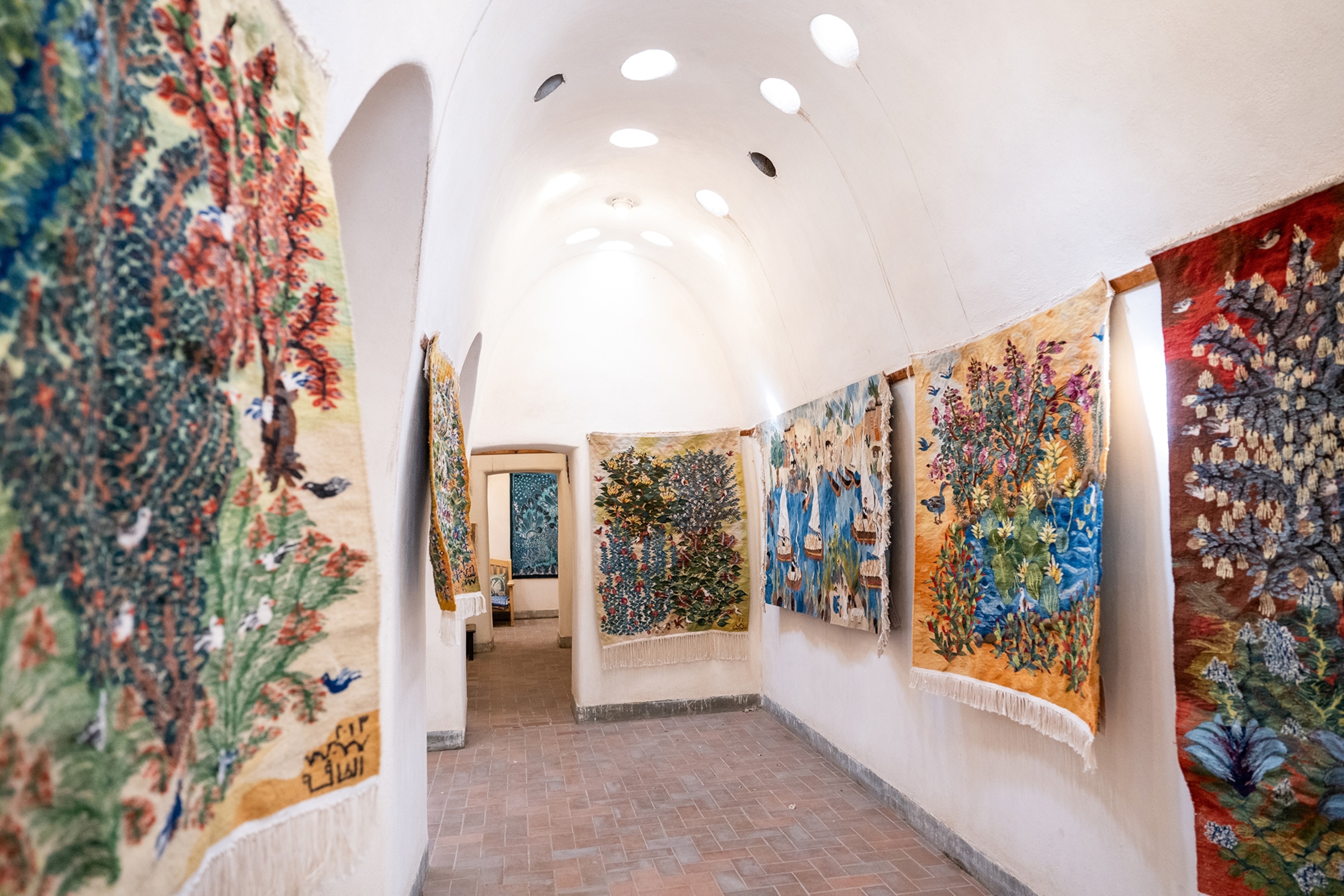 The interior of a simple gallery space with arched stone walls and rugs on display.