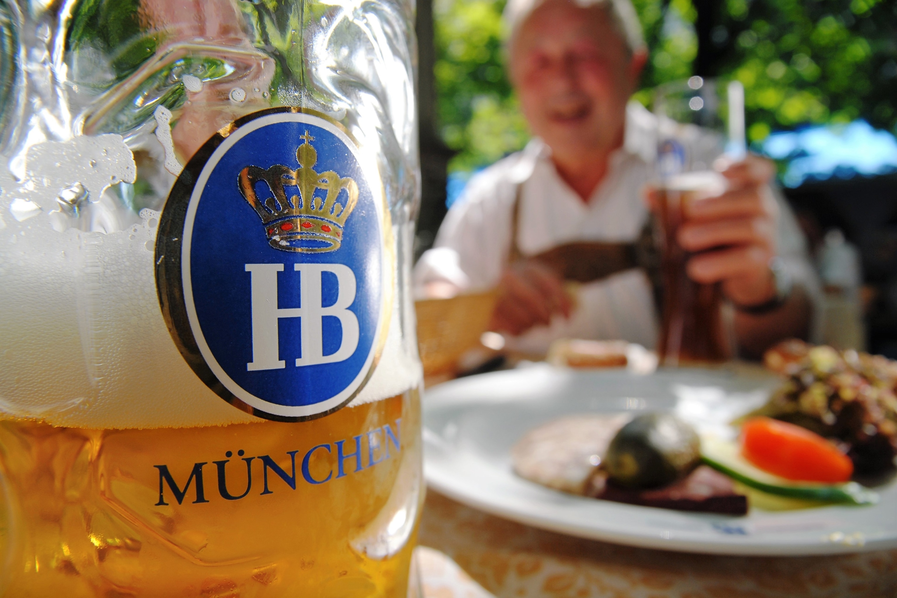 A dynamic close-up of a beer glass on an outdoor beer bench with a blurred man in lederhosen and a plate of traditional food in the background.