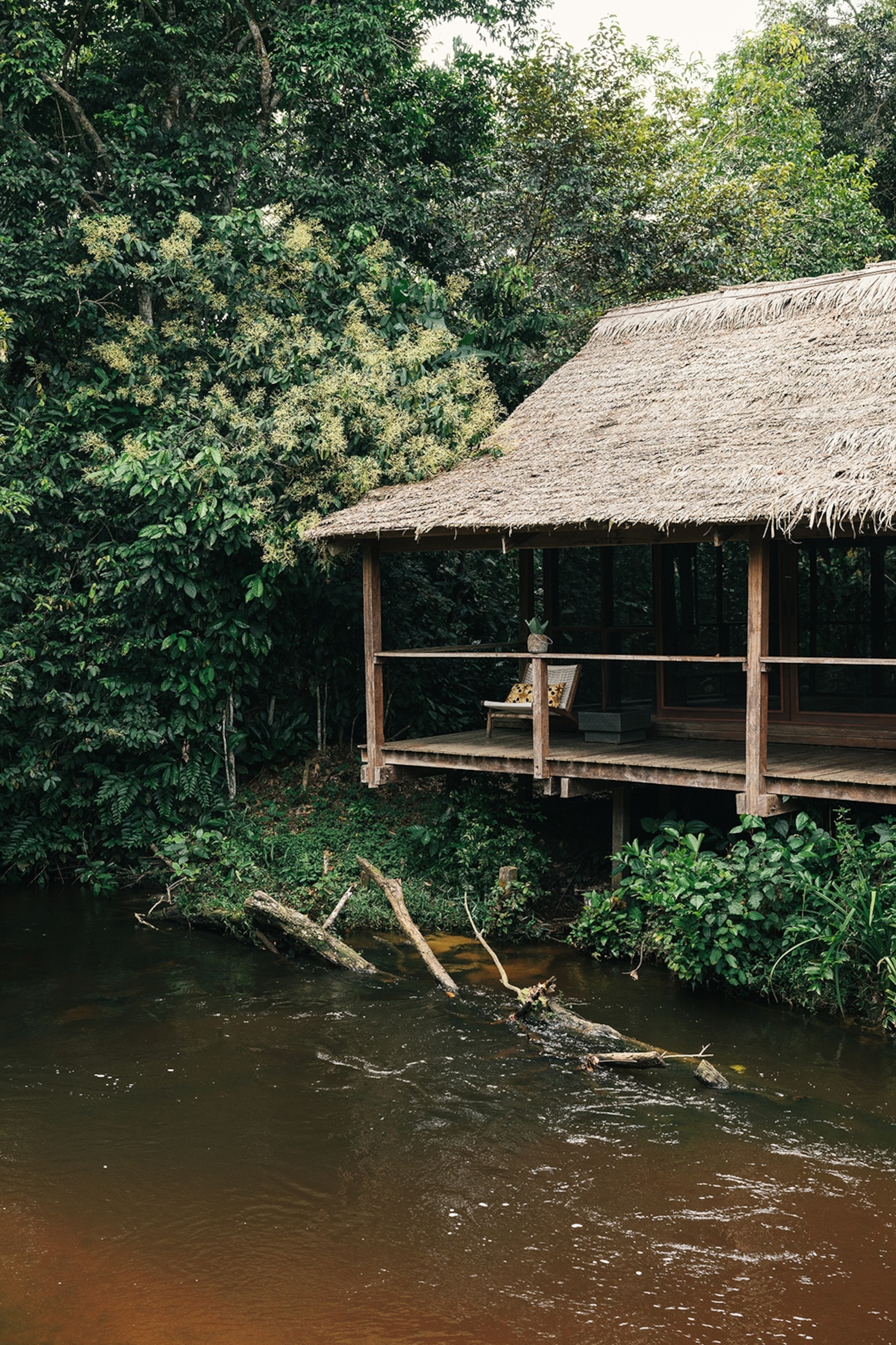 A simple, wooden lodge in the middle of a rainforest with overhanging balcony as a river courses past.