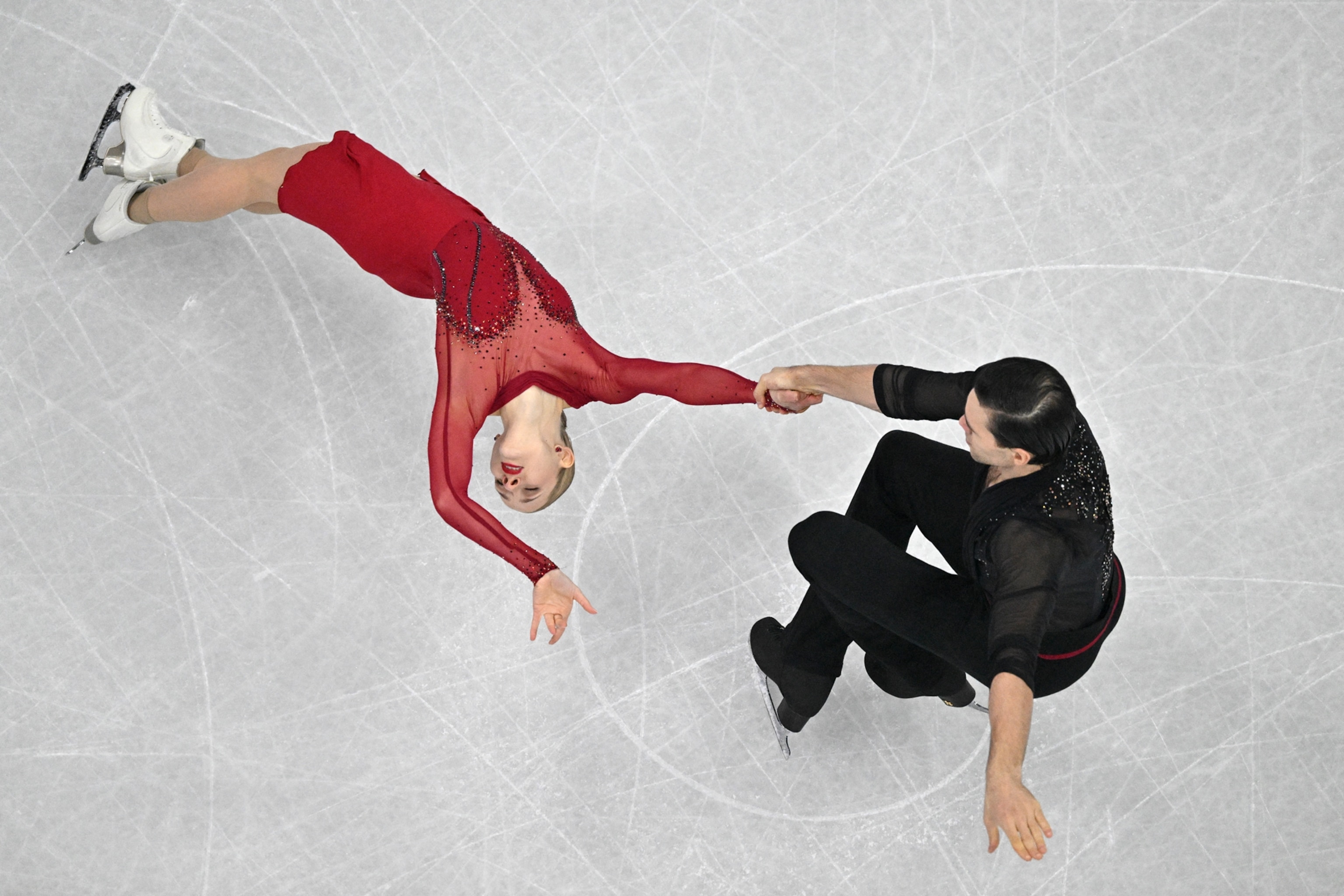 A woman in red leans back as she glides on the ice with a partner.