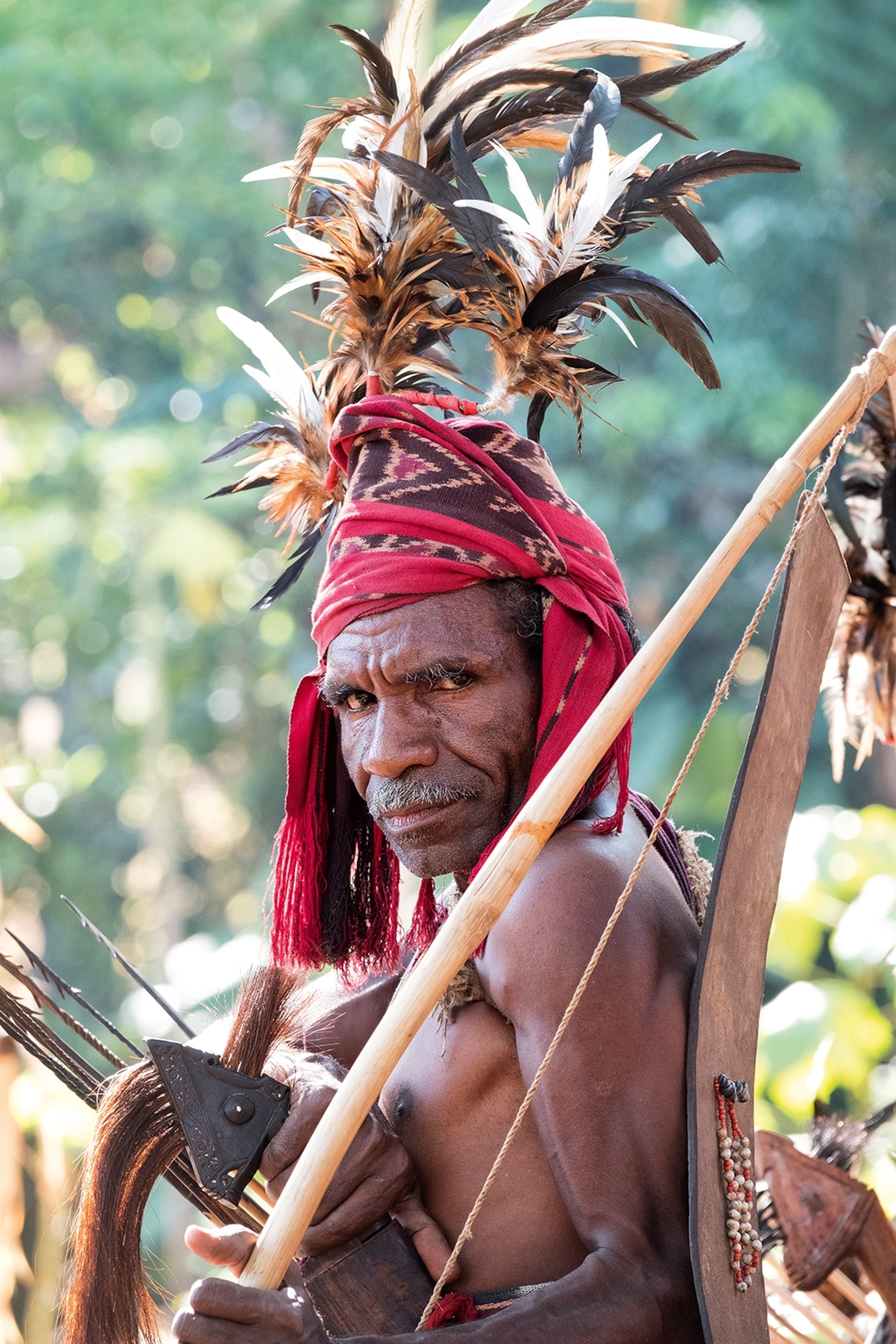 An elderly, local man wearing a feathered headdress and cross and bow, looking directly into the camera.