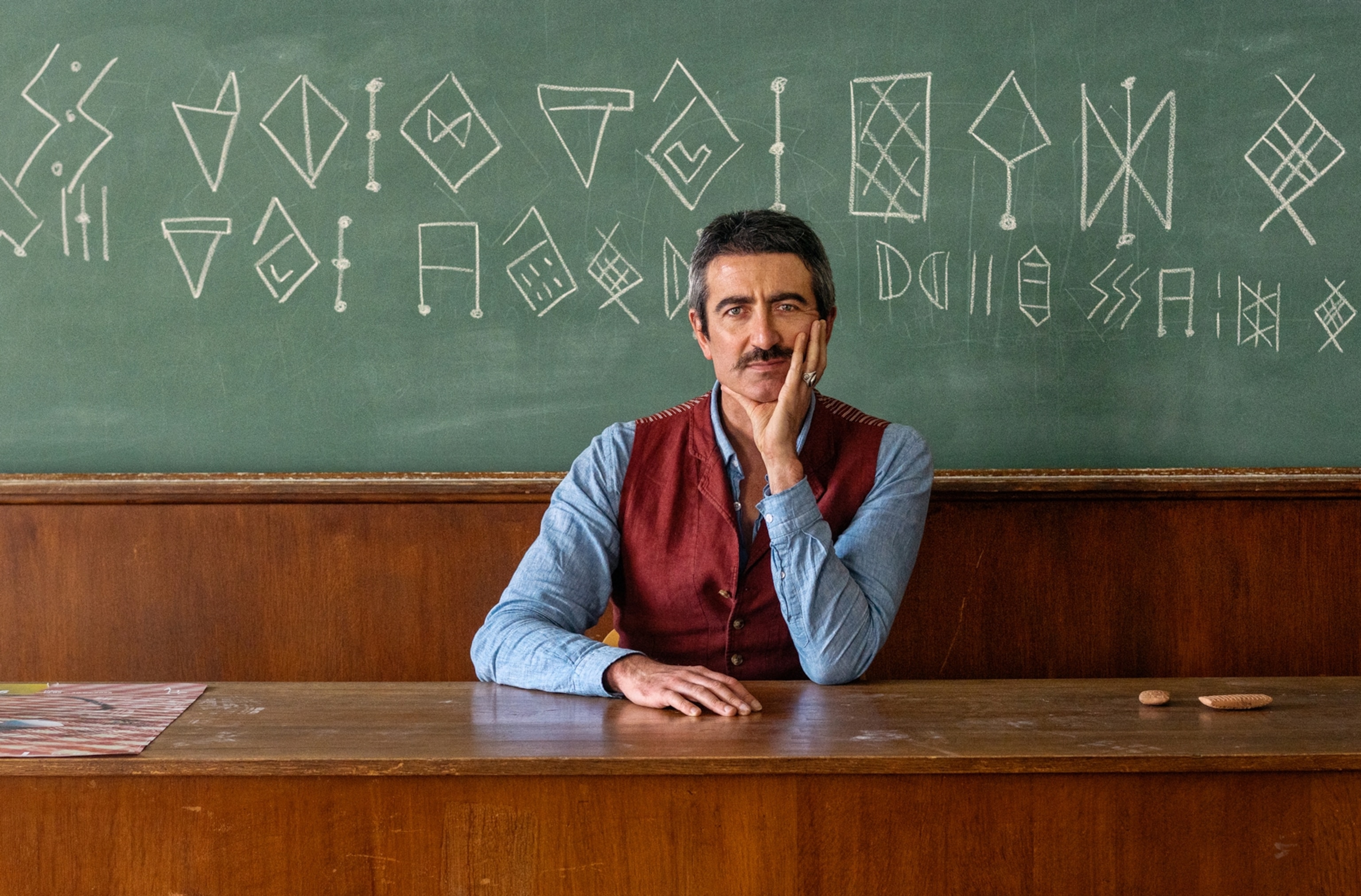 A man sits for a portrait at a desk in front of a chalkboard that has characters written on it.