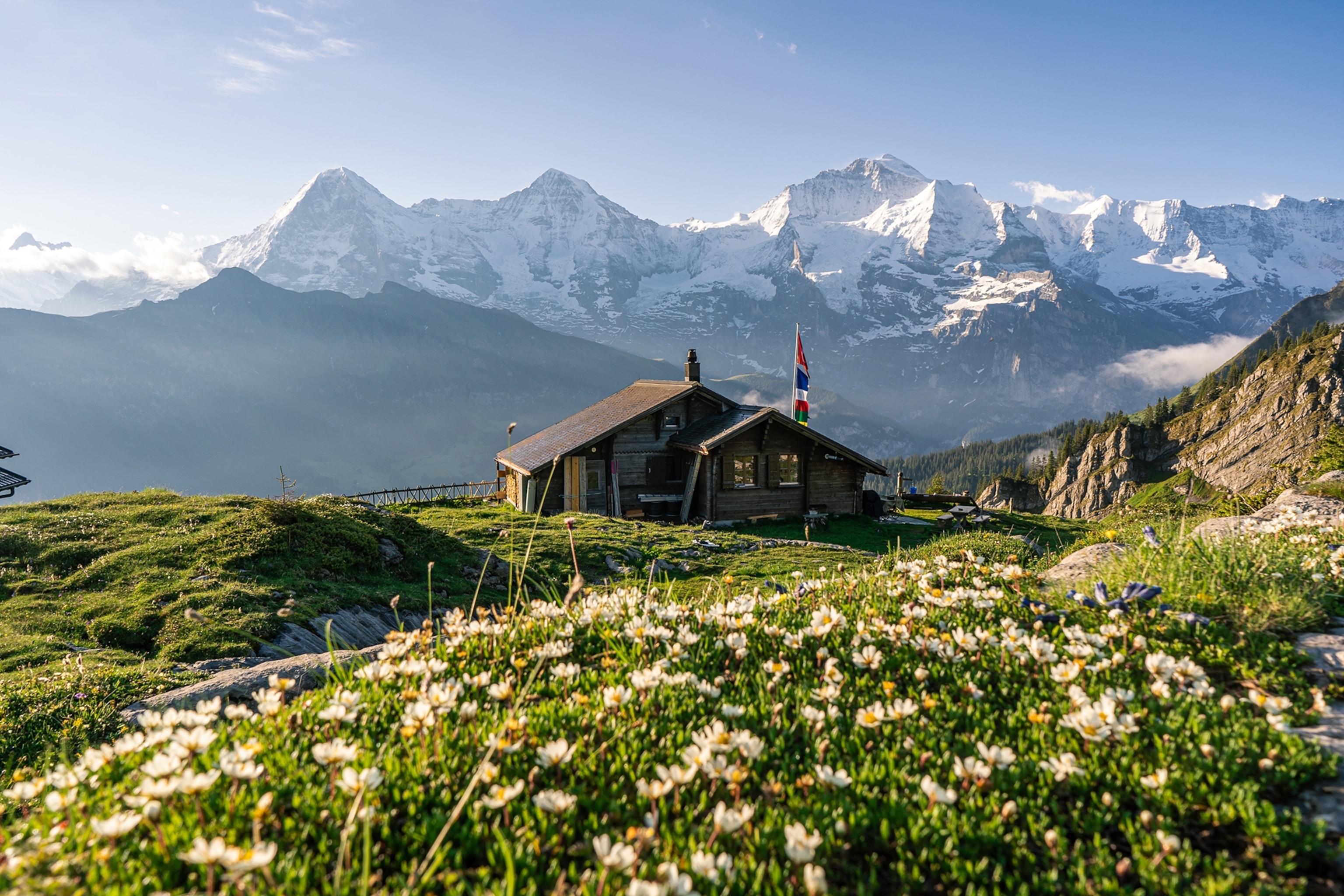 A wooden hut stands in a green meadow in front of large snow-capped peaks.