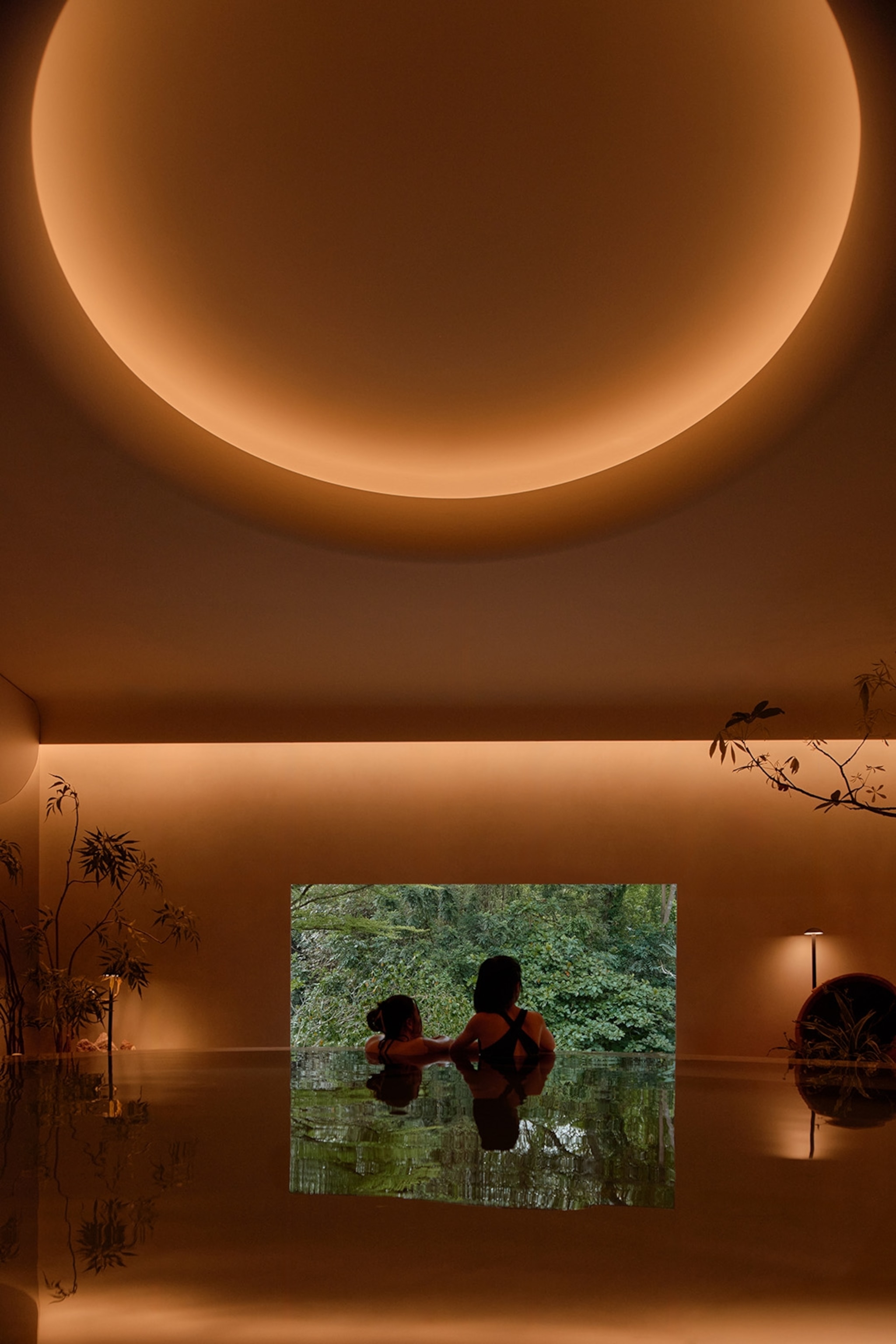 Two guests leaning on the edge of a round indoor spa pool looking out through a square window into a garden.