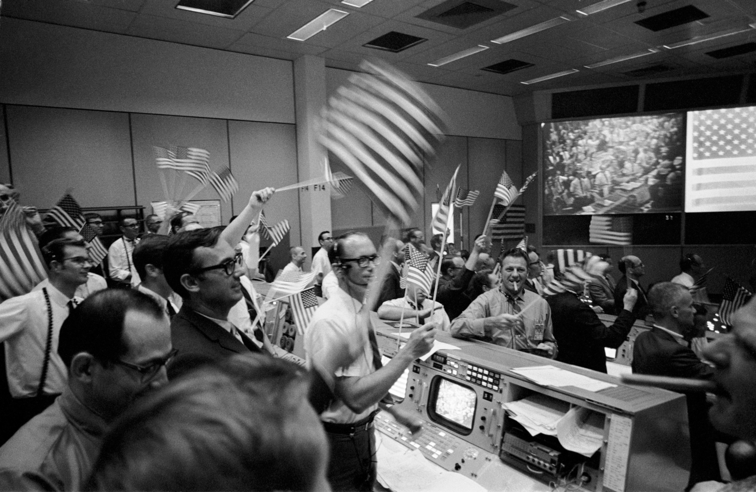 Men standing inside the Mission Control Center waving USA flags and celebrating.