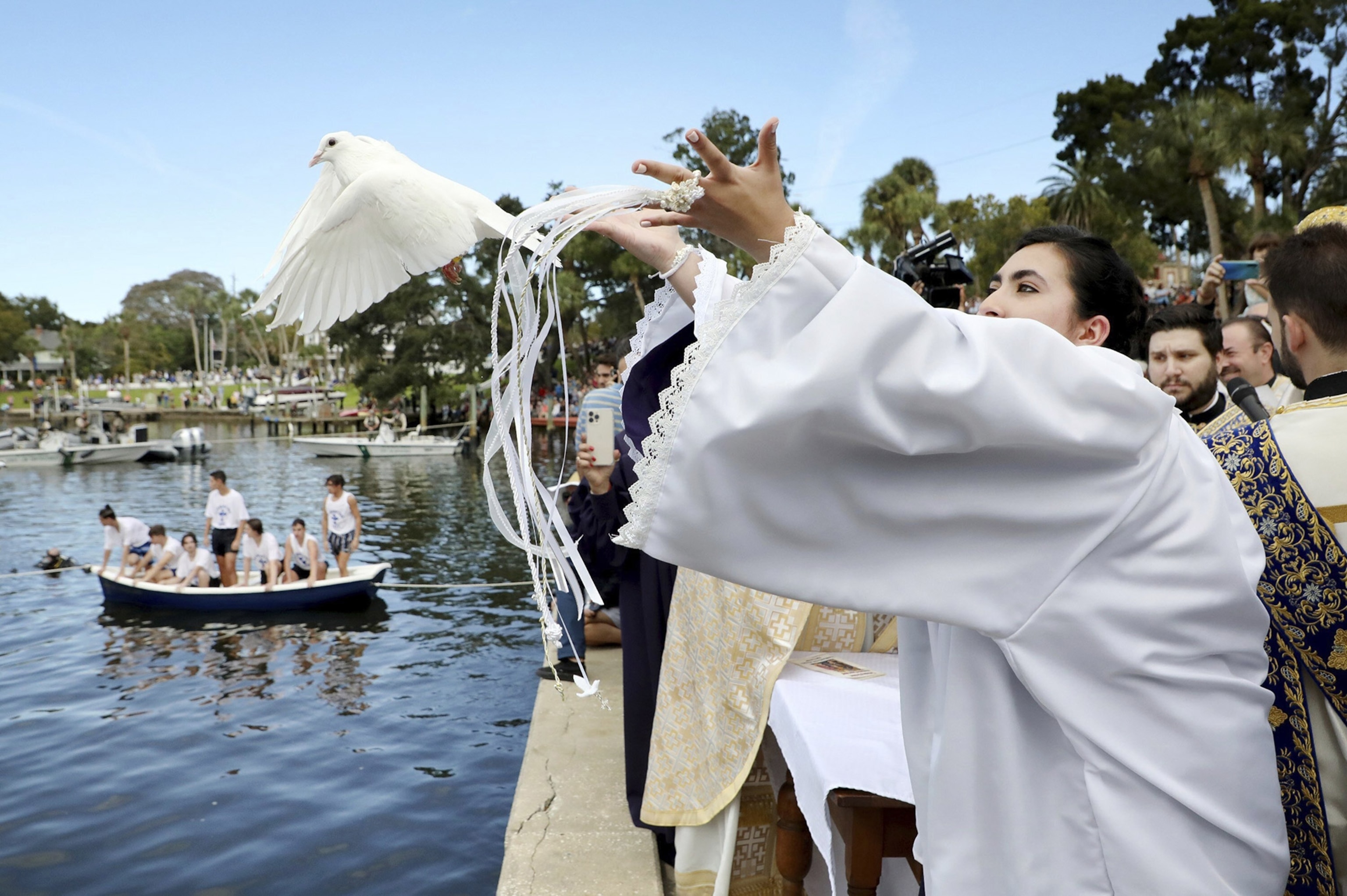 A girl in a white robe releases a white dover over the water, in the background a group of people in white t-shirts sit in a boat