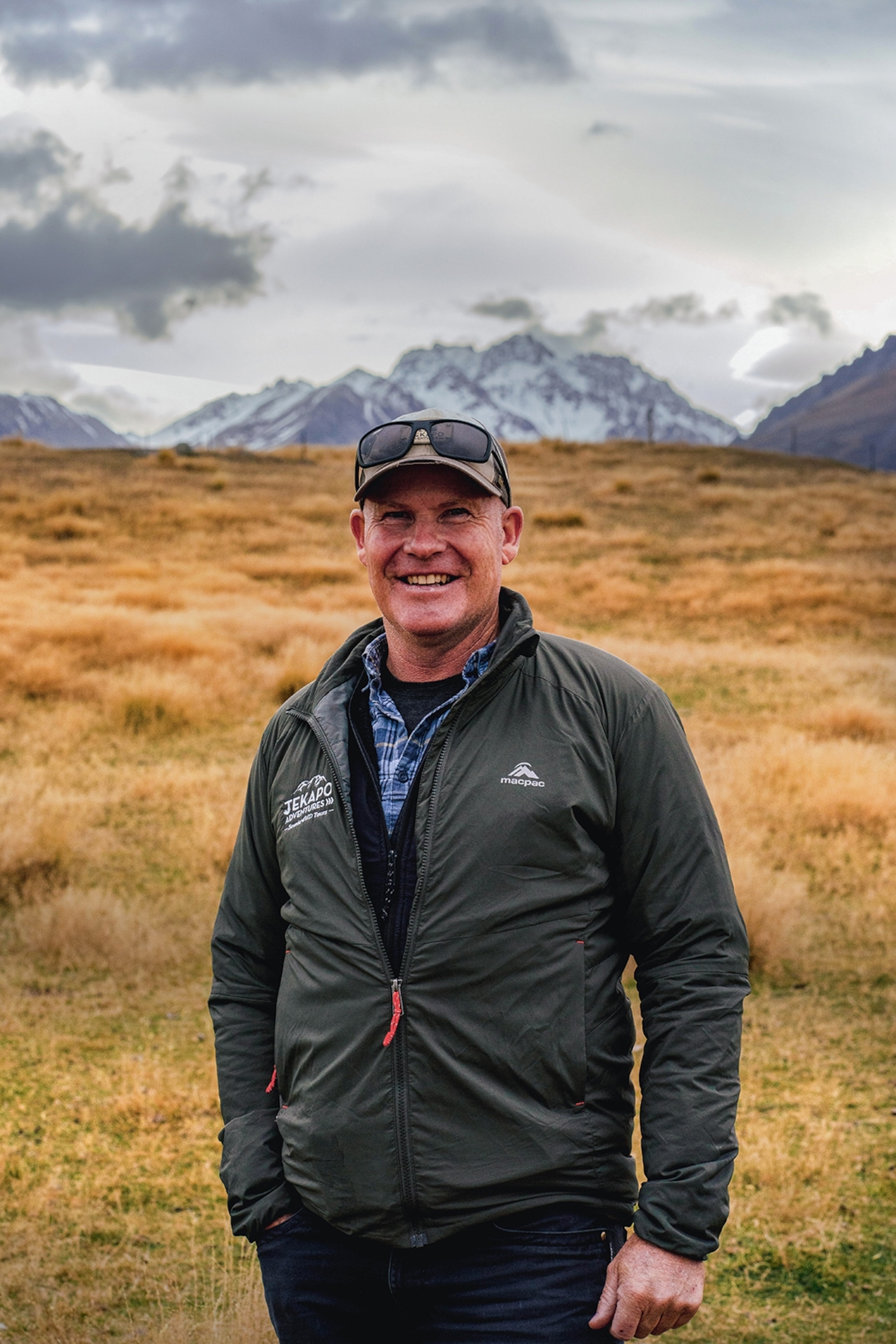 A friendly man wearing outdoor gear and a cap, posing for a portrait with a mountaineous landscape in the back.