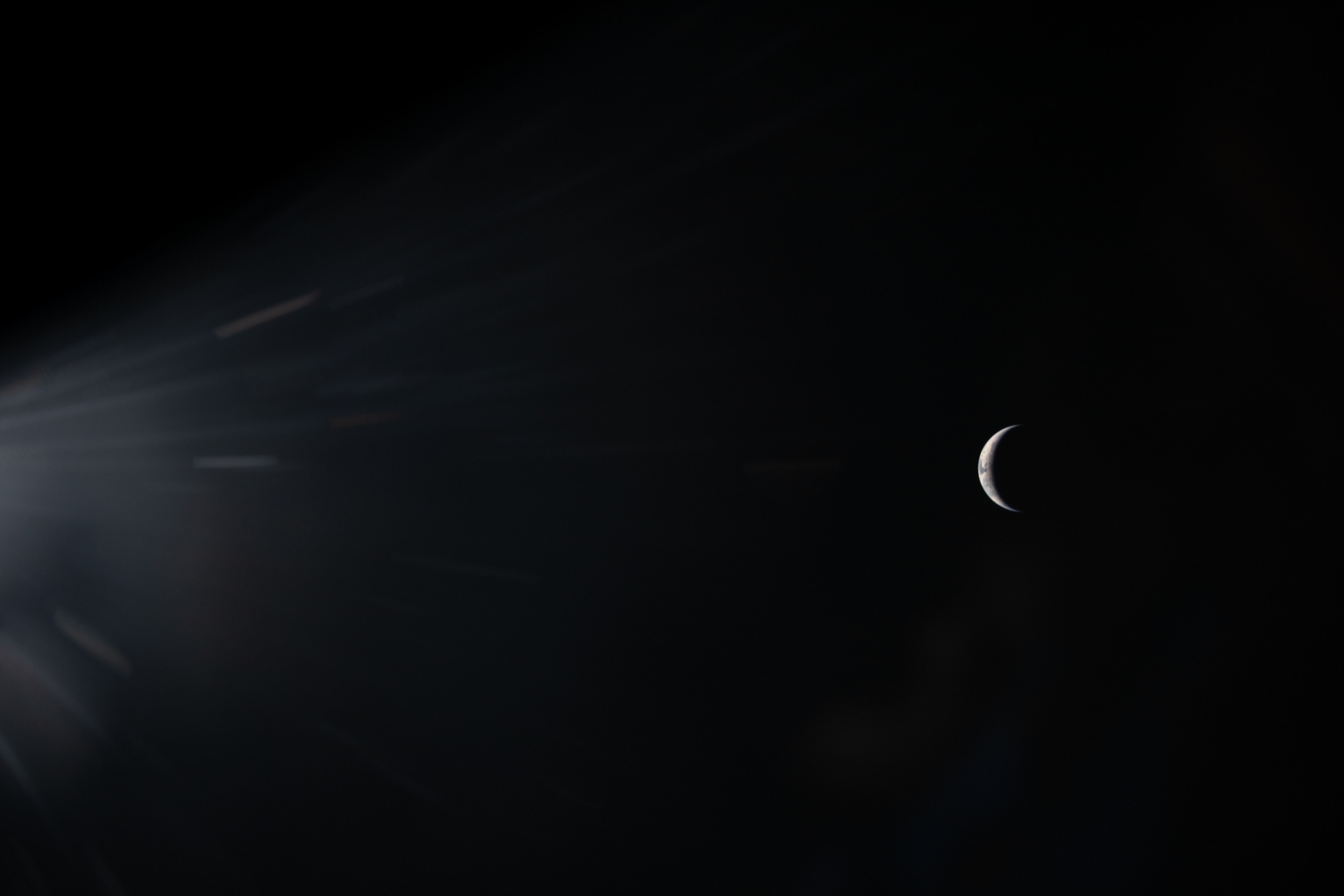 A thin crescent seen against a dark background with radiant light beams coming from the left
