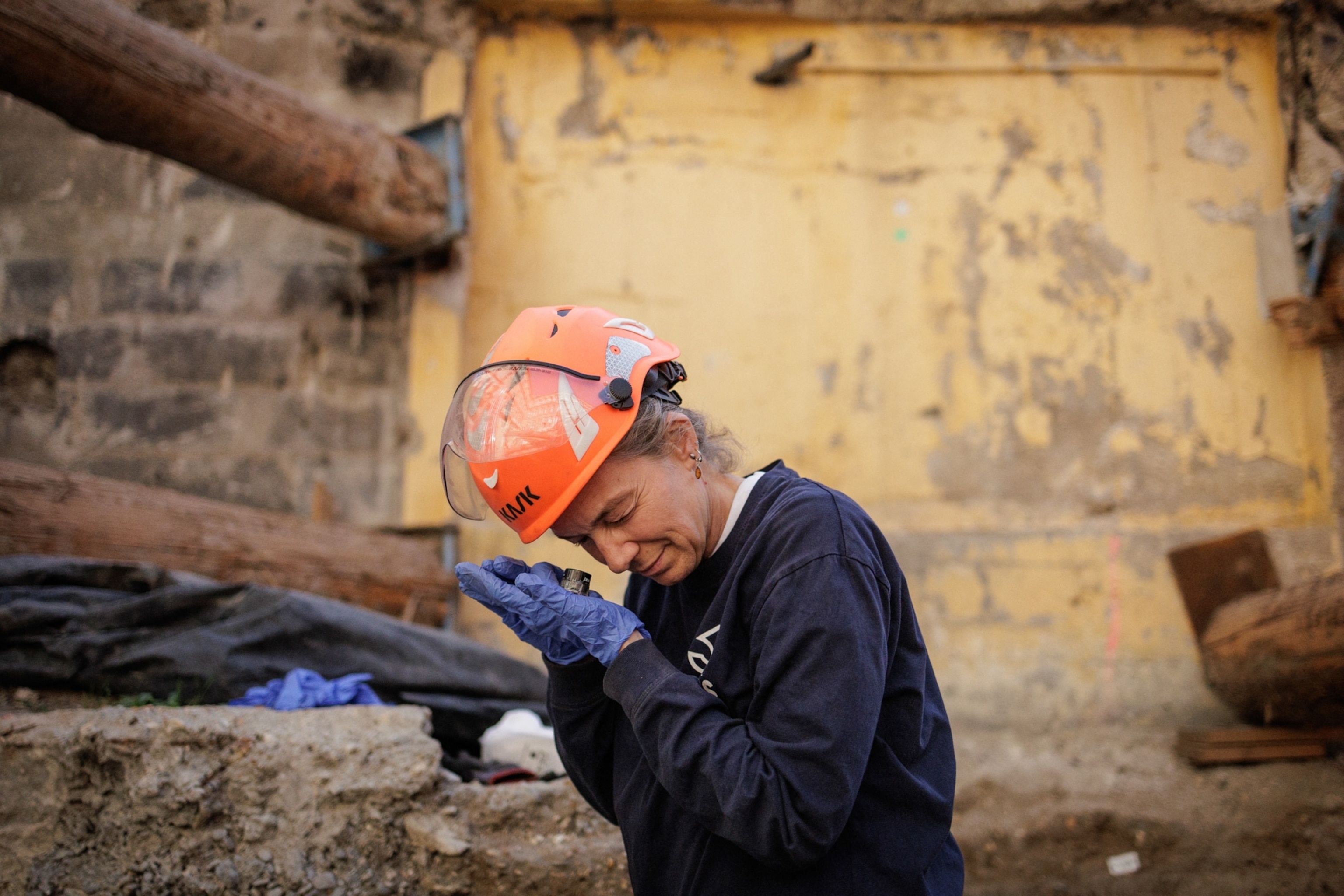 An archaeologist wearing a safety helmet and gloves carefully examines a small artifact at an excavation site surrounded by exposed stone walls and earth.