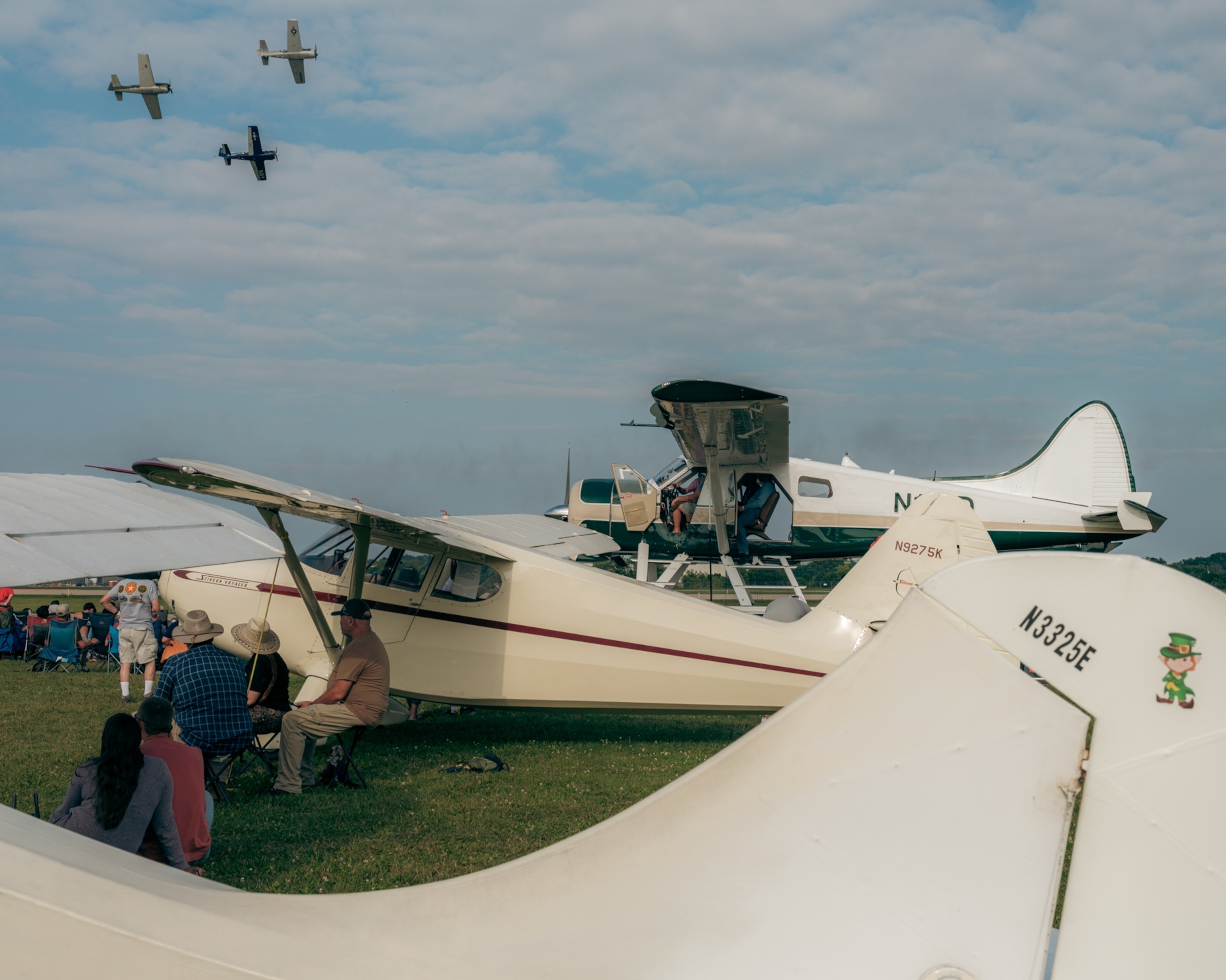 A group of vintage planes are parked on a grassy field under a cloudy blue sky. Spectators sit nearby, watching three planes flying overhead