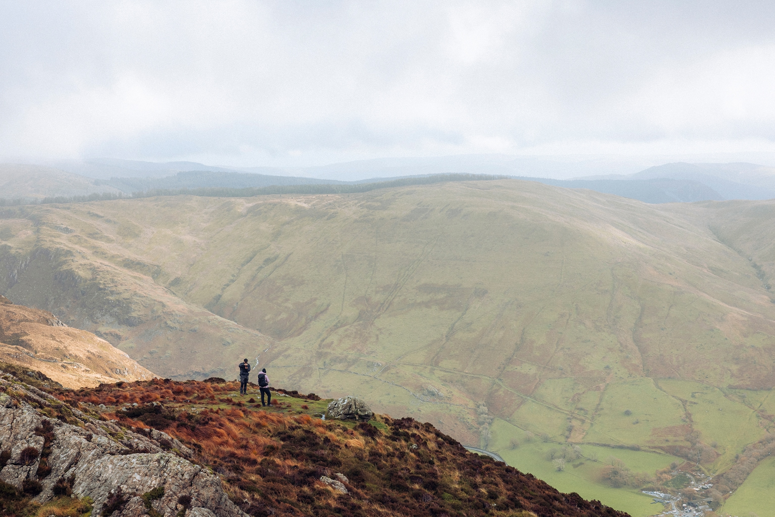 Two hikers standing on the cliff edge of a mountain with a misty landscape in the background.