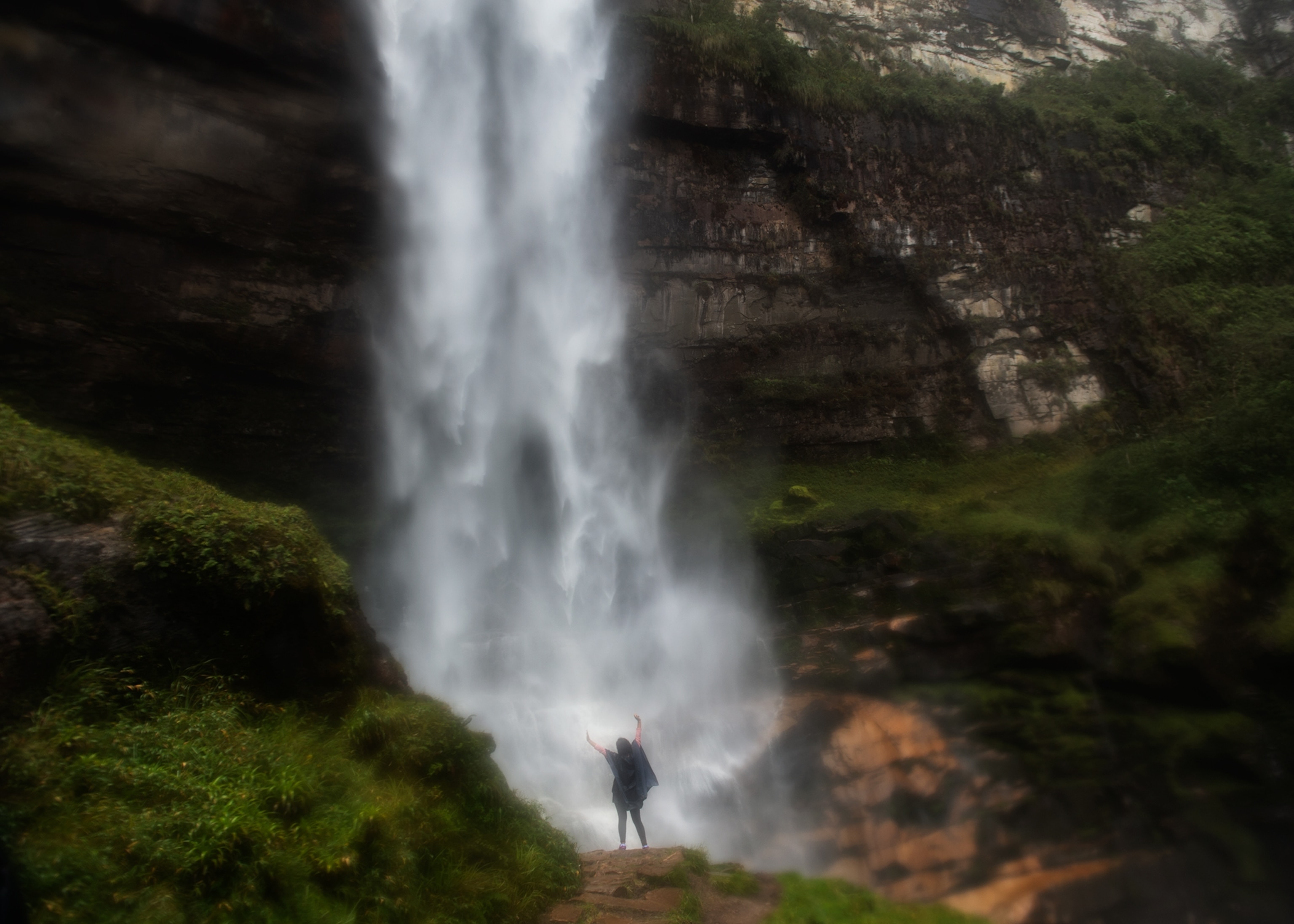 A person stands with raised arms at the base of a powerful waterfall, surrounded by steep rocky cliffs and lush green vegetation.