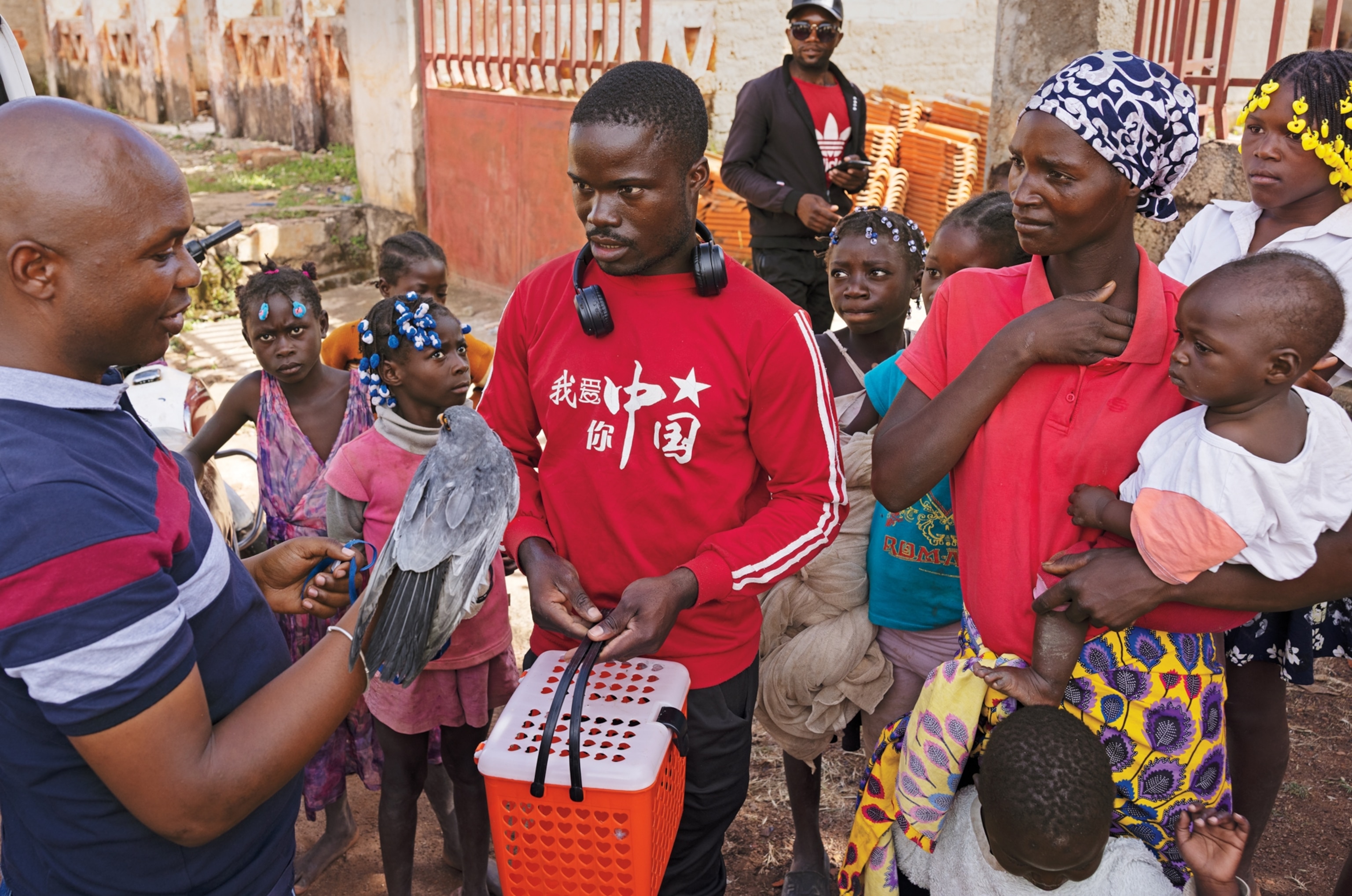 Man and woman in red shirts surrounded by children listening to a man holding a grey bird.