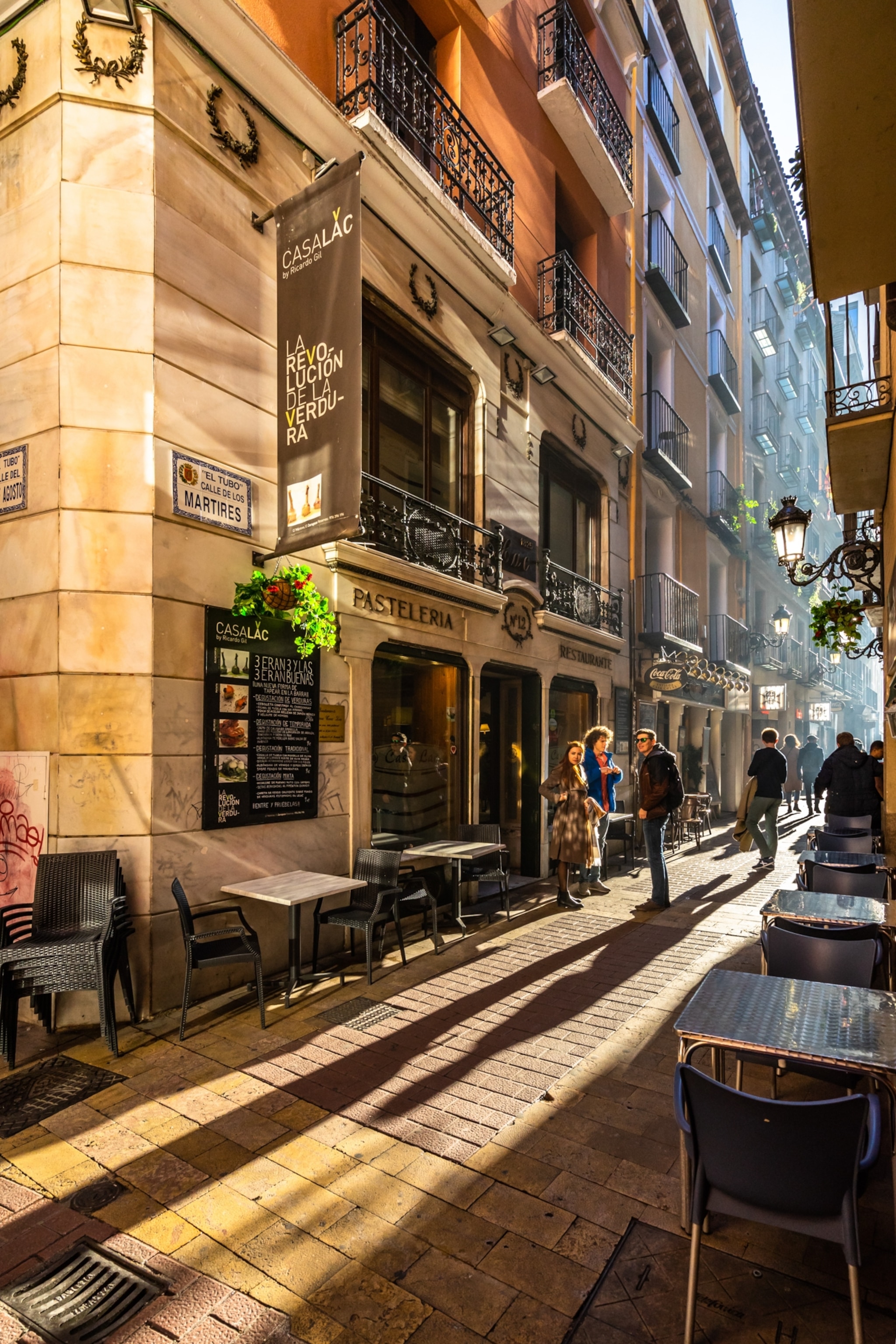 A tight street has light streaming through elongating the shadows cast of the people standing.