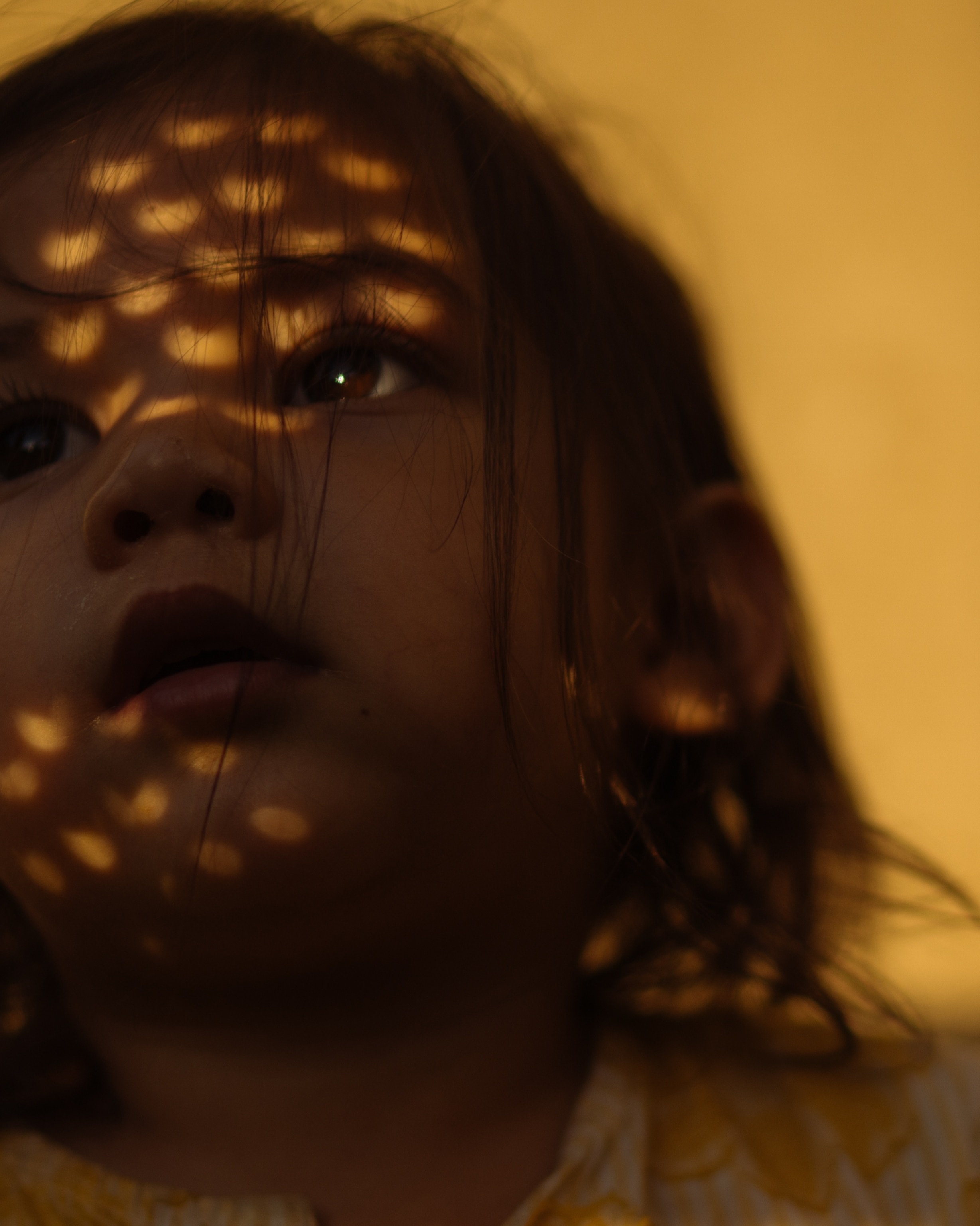 Close-up portrait of a child gazing upward in warm amber light, with patterned sunlight during a partial eclipse casting glowing spots resembling hearts across their face and tousled hair