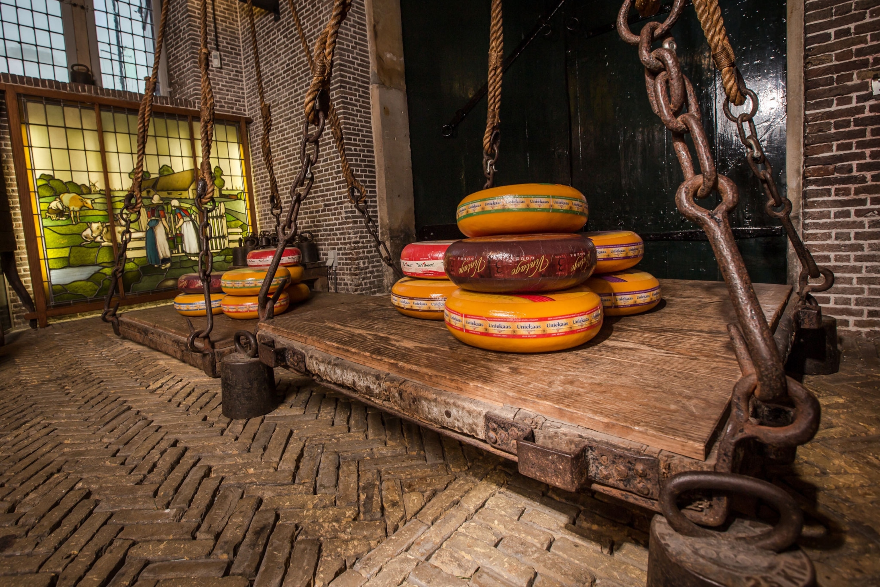 Large wheels of cheese sit atop old wooden platforms, suspended by heavy chains in a brick-walled factory.