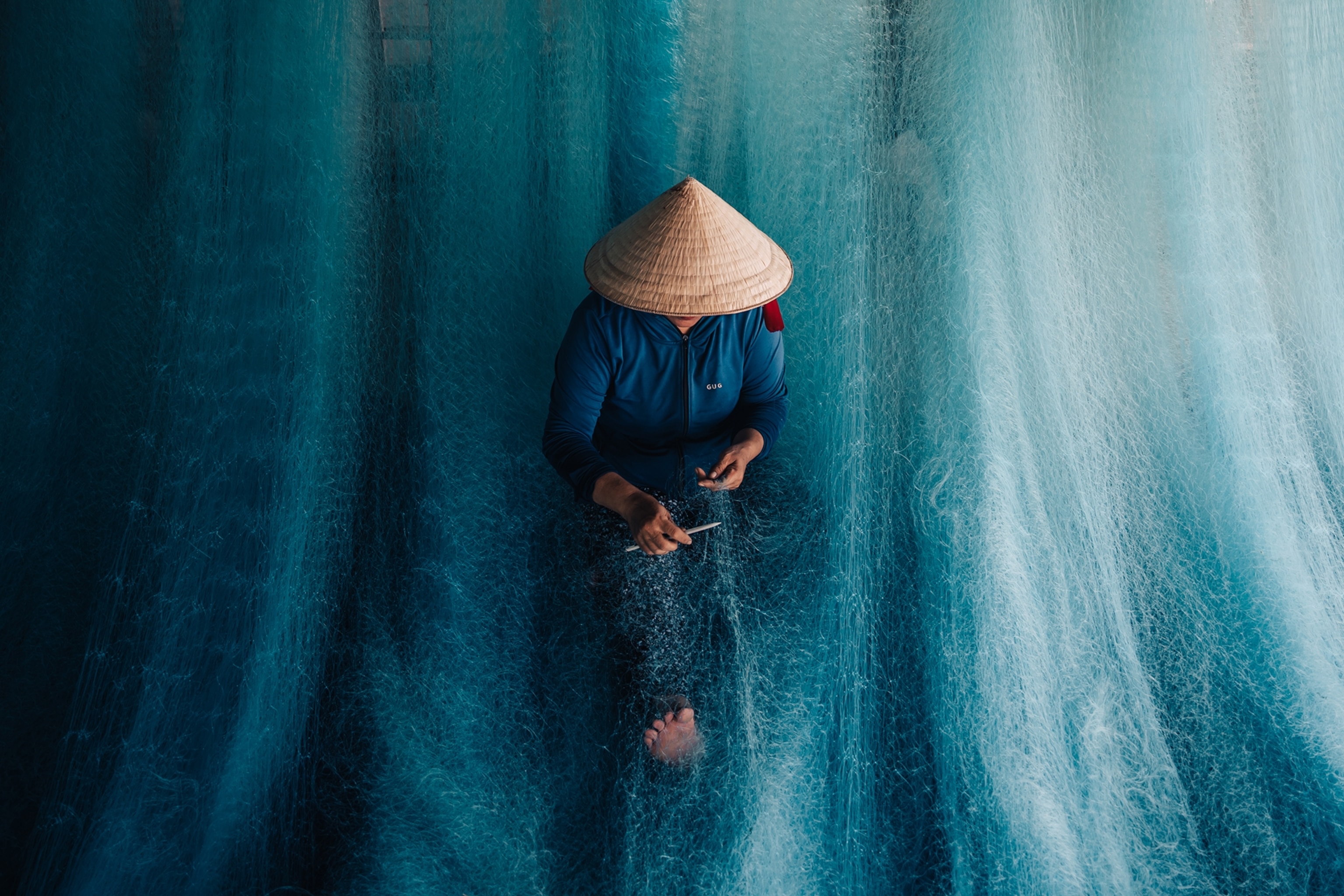 A Vietnamese fisher wearing a straw, cone hat and sitting in the middle of a fishing net.
