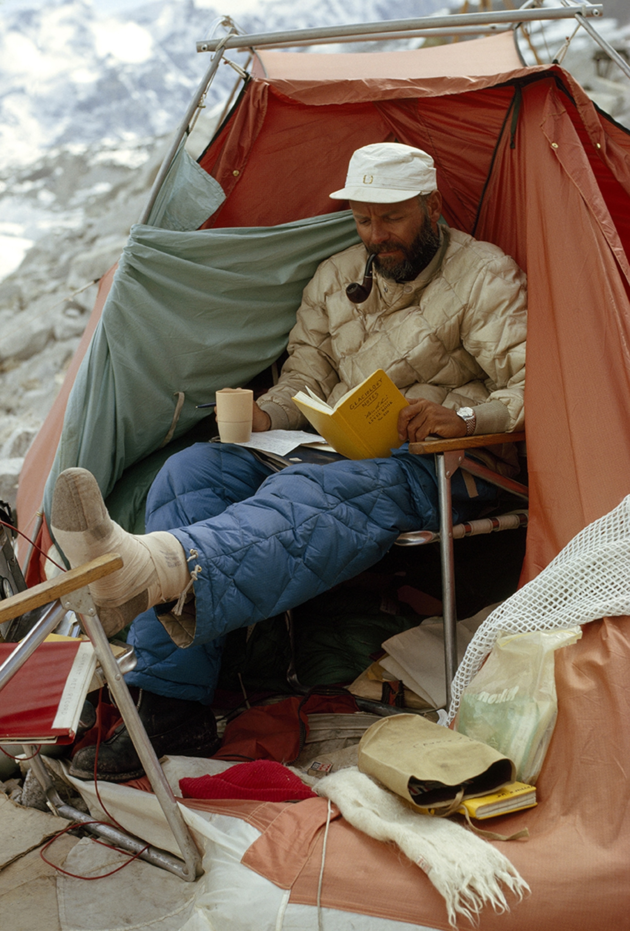 Foot broken by tumbling boulder, glaciologist Maynard M. Miller keeps up his work from his tented domain on “Nob Hill,” in Base Camp. He melted snow samples of previous years to measure the content of tritium, pollen, dust, salts, and microorganisms. When West Ridge climbers Hornbein and Unsoeld came into camp desperate for liquids, Miller gave them a precious sample of water from 12-year-old ice taken from crevasse wall.