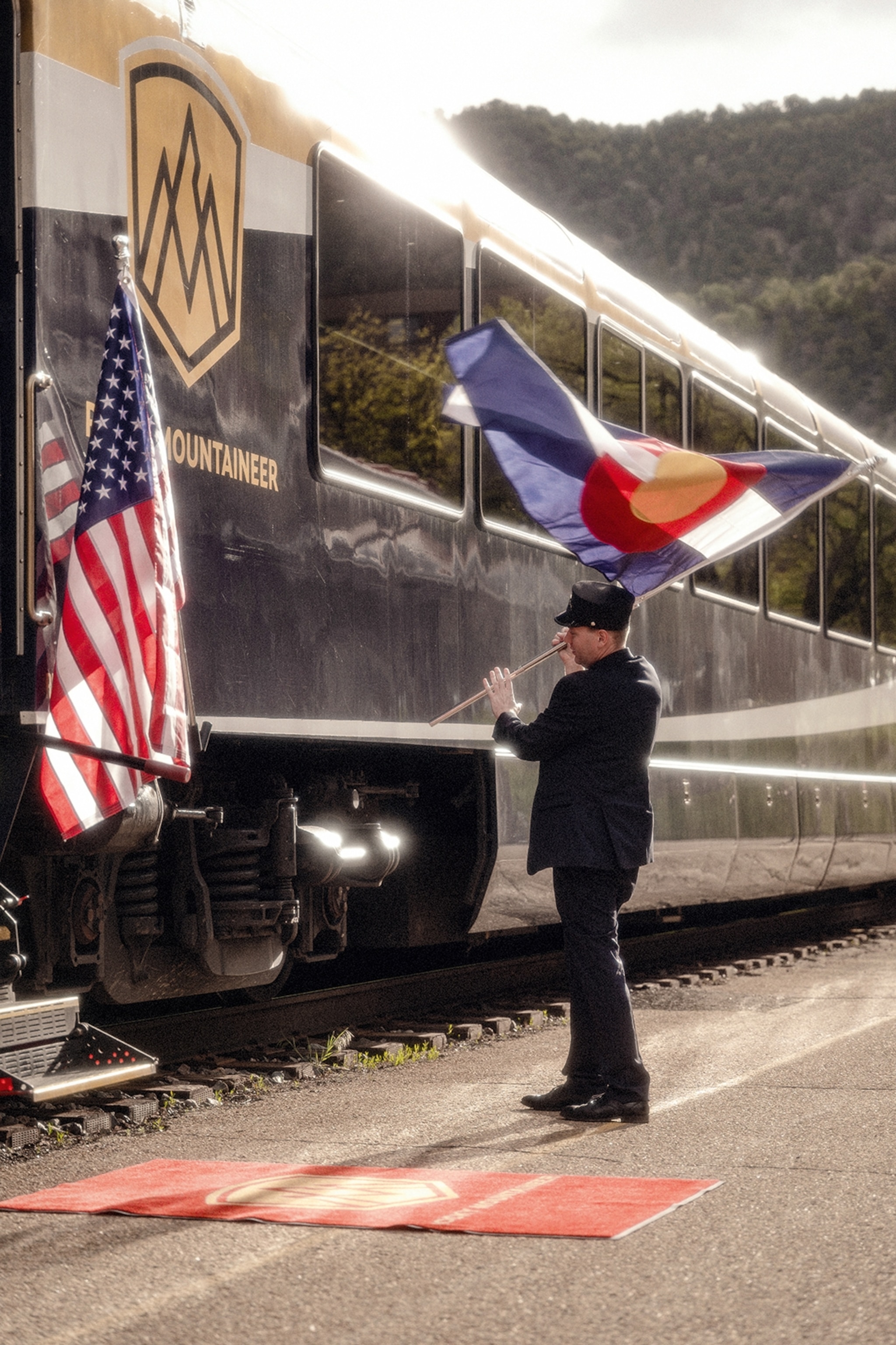 A shiny train halted at a train station with a serviceman holding an American flag by one of the train's doors, a red carpet is rolled out below.