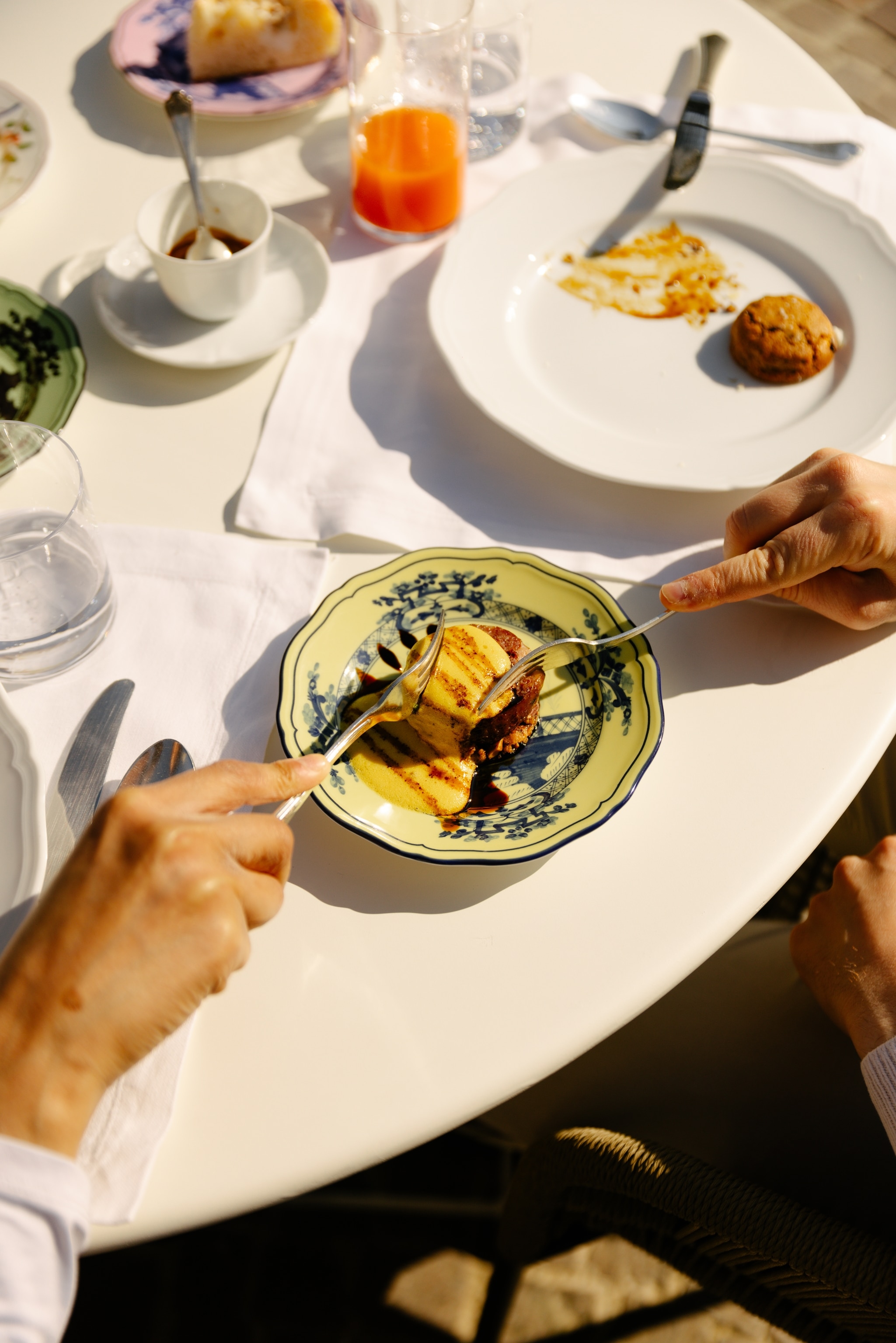 A plate on an outdoor table with two hands digging into the food with forks.