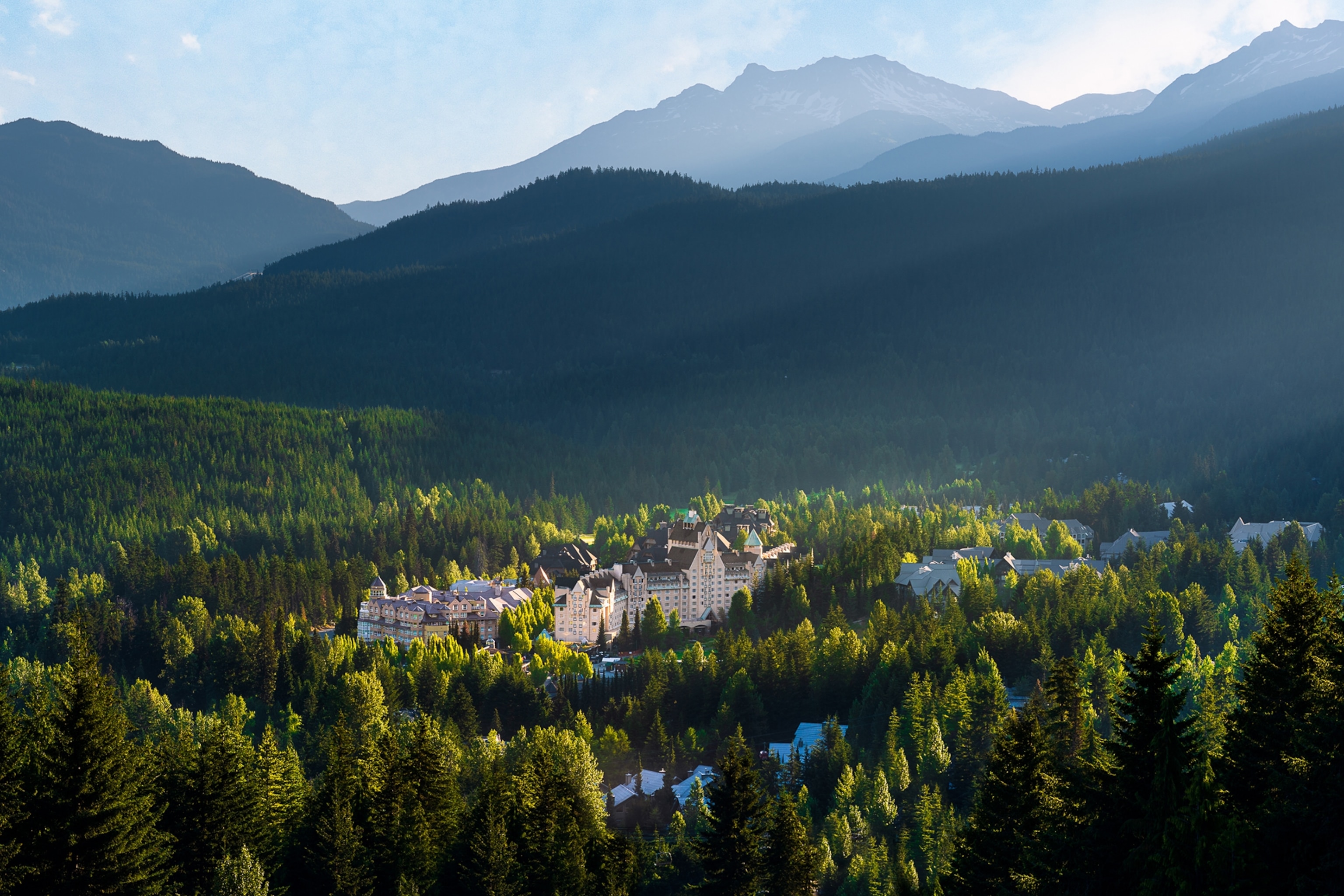 A castle nestled into a pine forested landscape with a mountain range in the background.