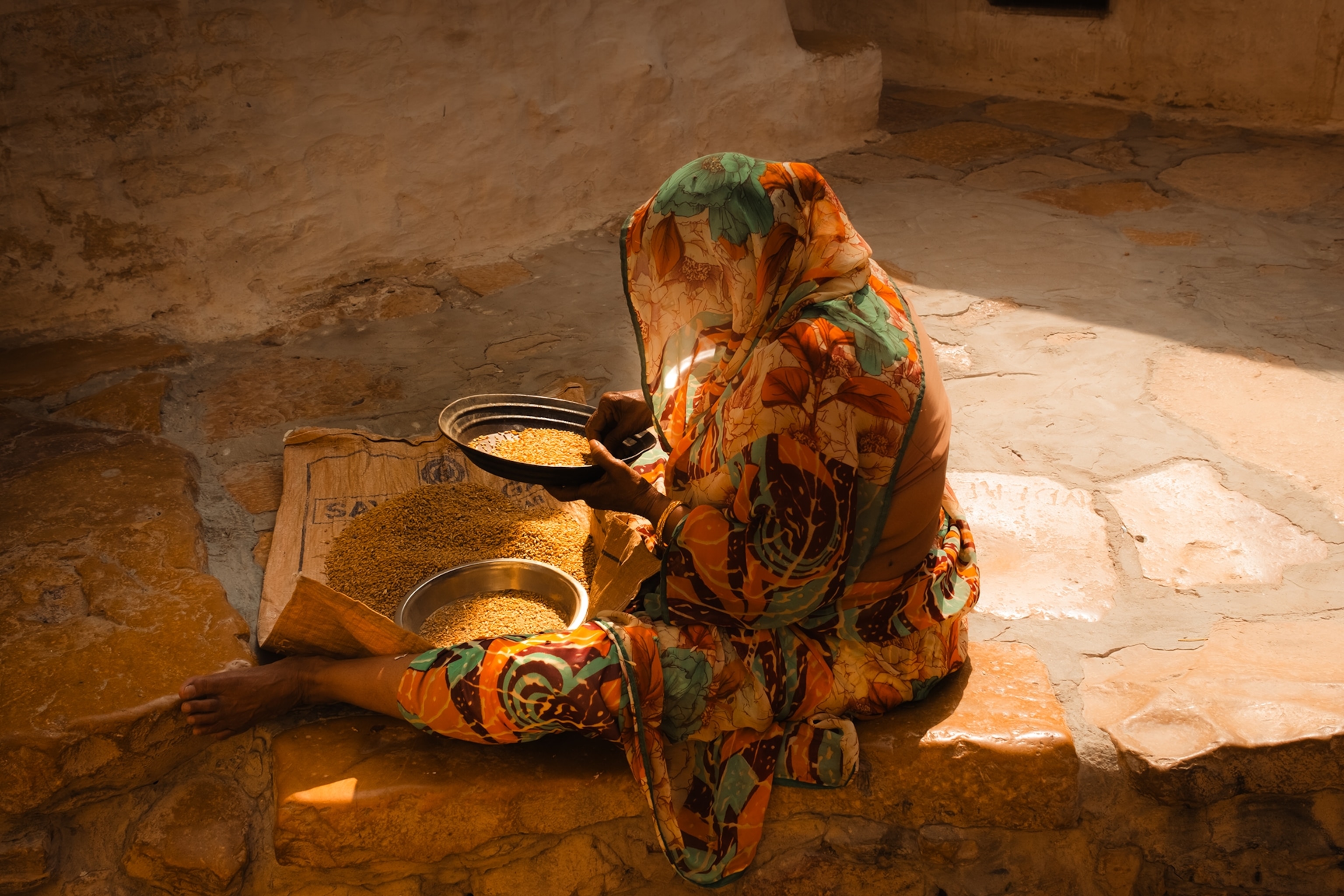 An Indian woman sitting on a stone floor in the shade, sieving lentils with her back to the camera.