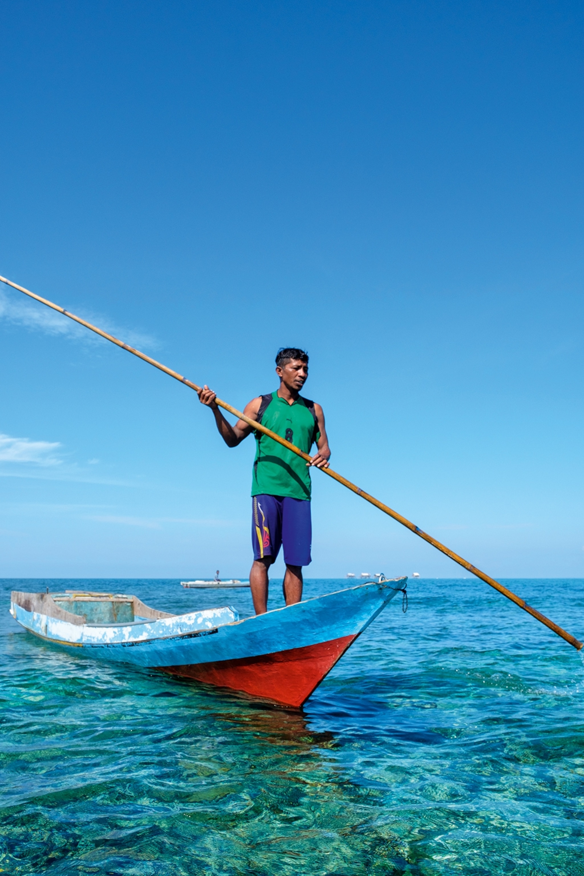 A local man in a simple tank top and shorts standing at the tip of a fishing boat, holding a spear in his hands.