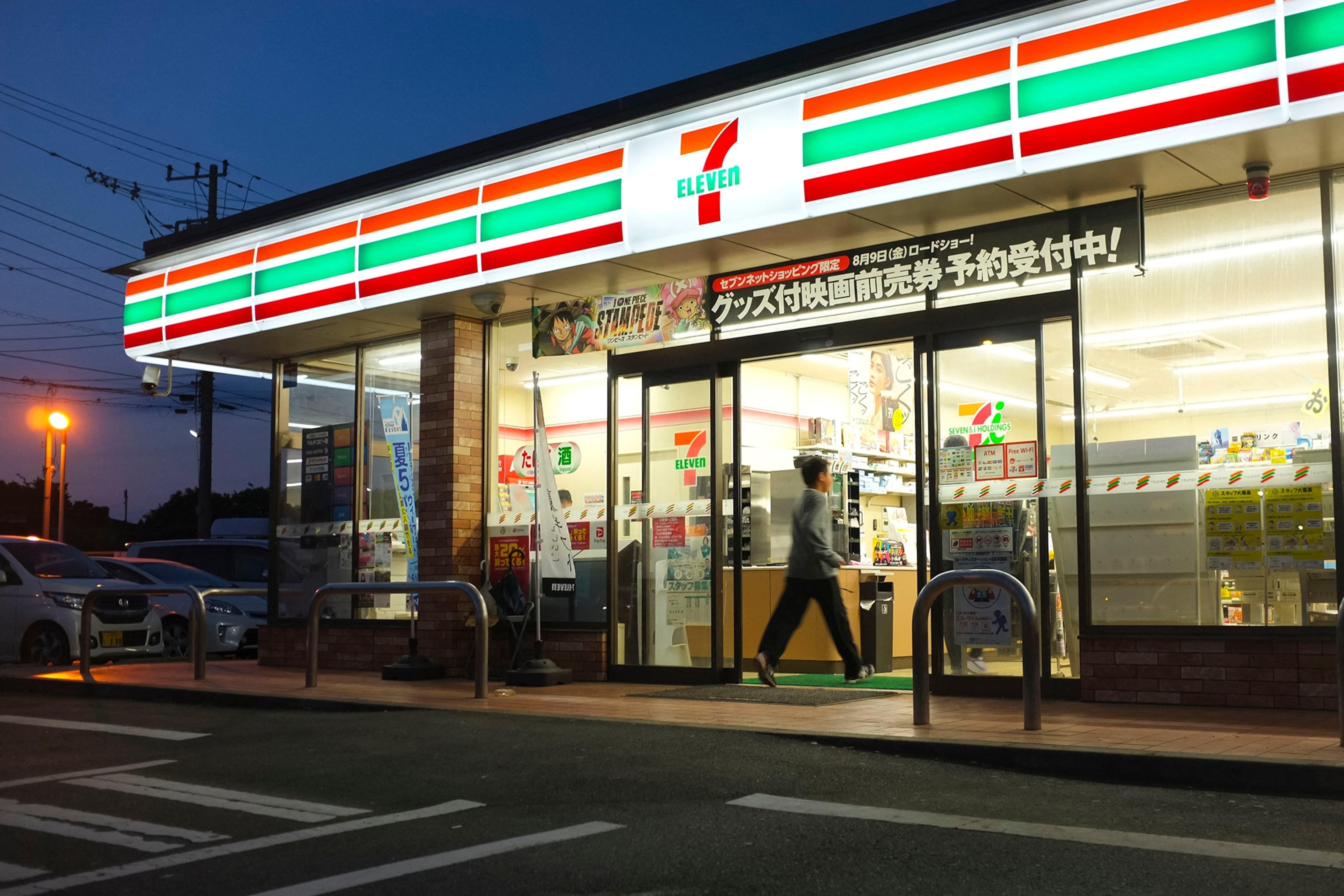 A 7-Eleven convenience store at night as a customer walks through the automatic, sliding doors.