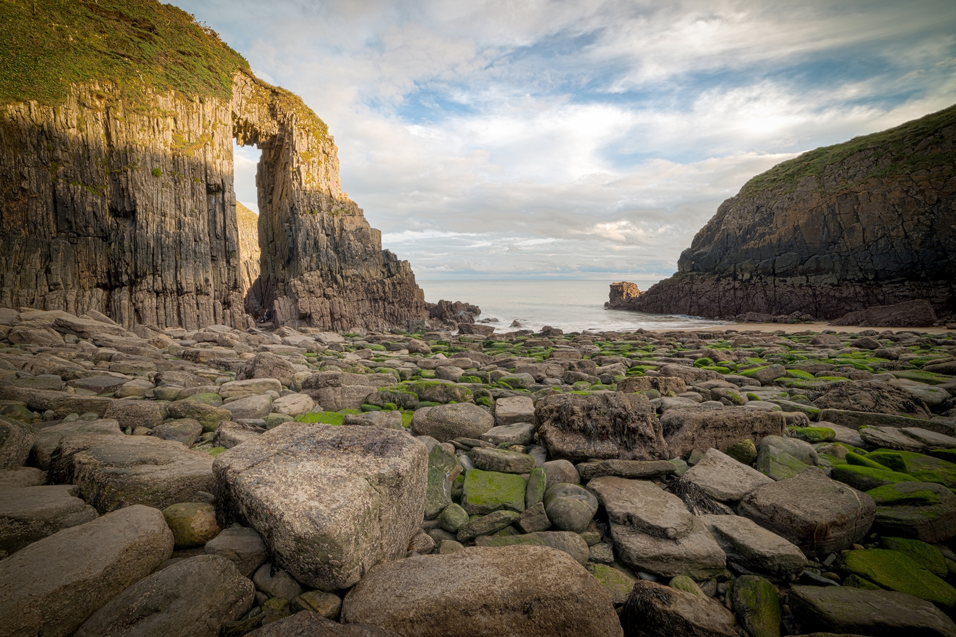 Rocky cove with tall archway on the left, headland on the right and the sea in the distance.