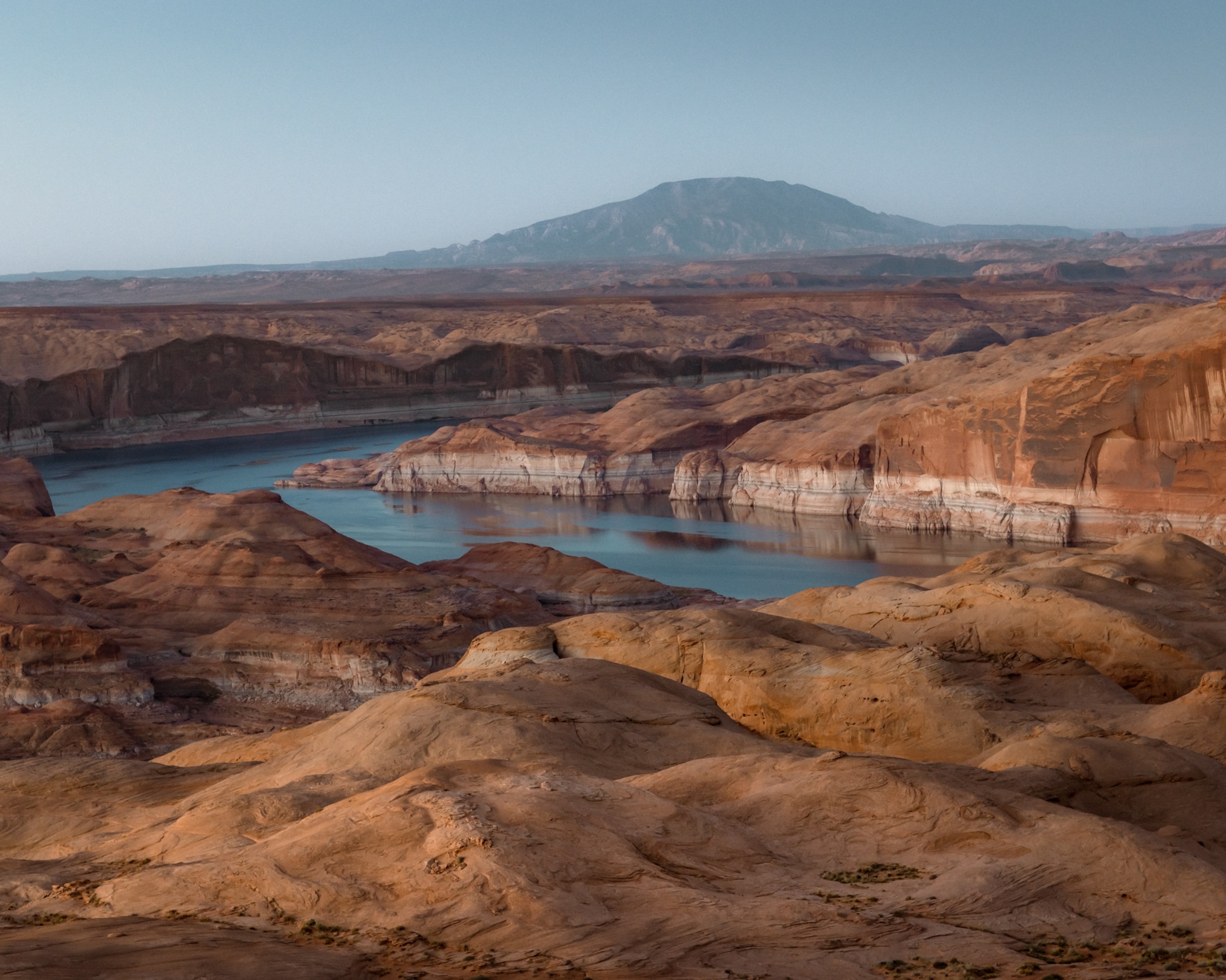 Desert landscape with red-orange rock formations surrounding a calm blue lake. The lake levels have receded, leaving behind a ring of white on the rocky walls.