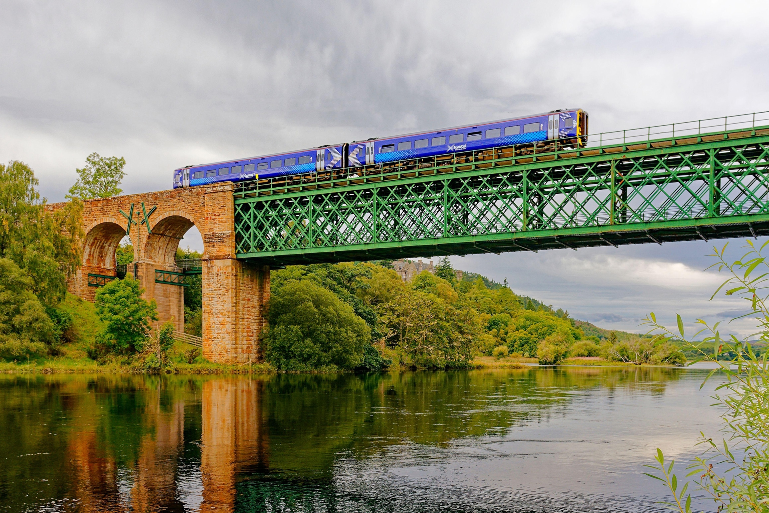 A blue train crosses a green, metal railway viaduct over a wide river - cloudy sky above.