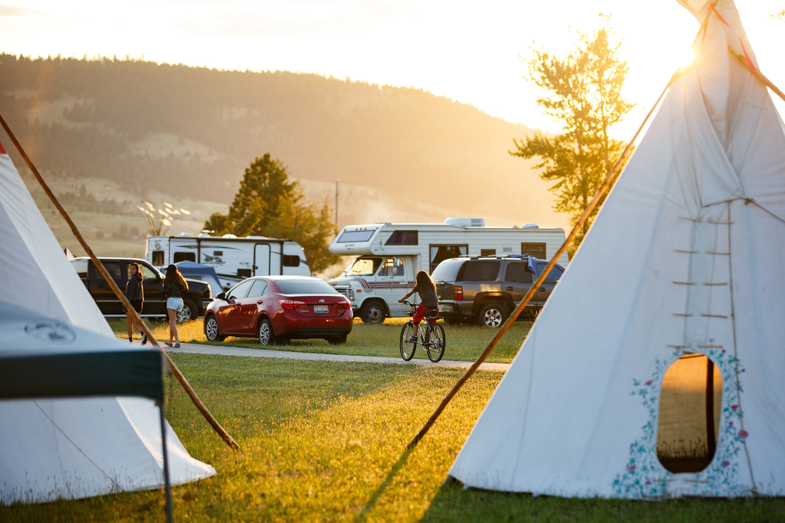 A camping site with tipi tents and parked campervans as the sun sets behind the horizon and a young girl drives by on a bike.