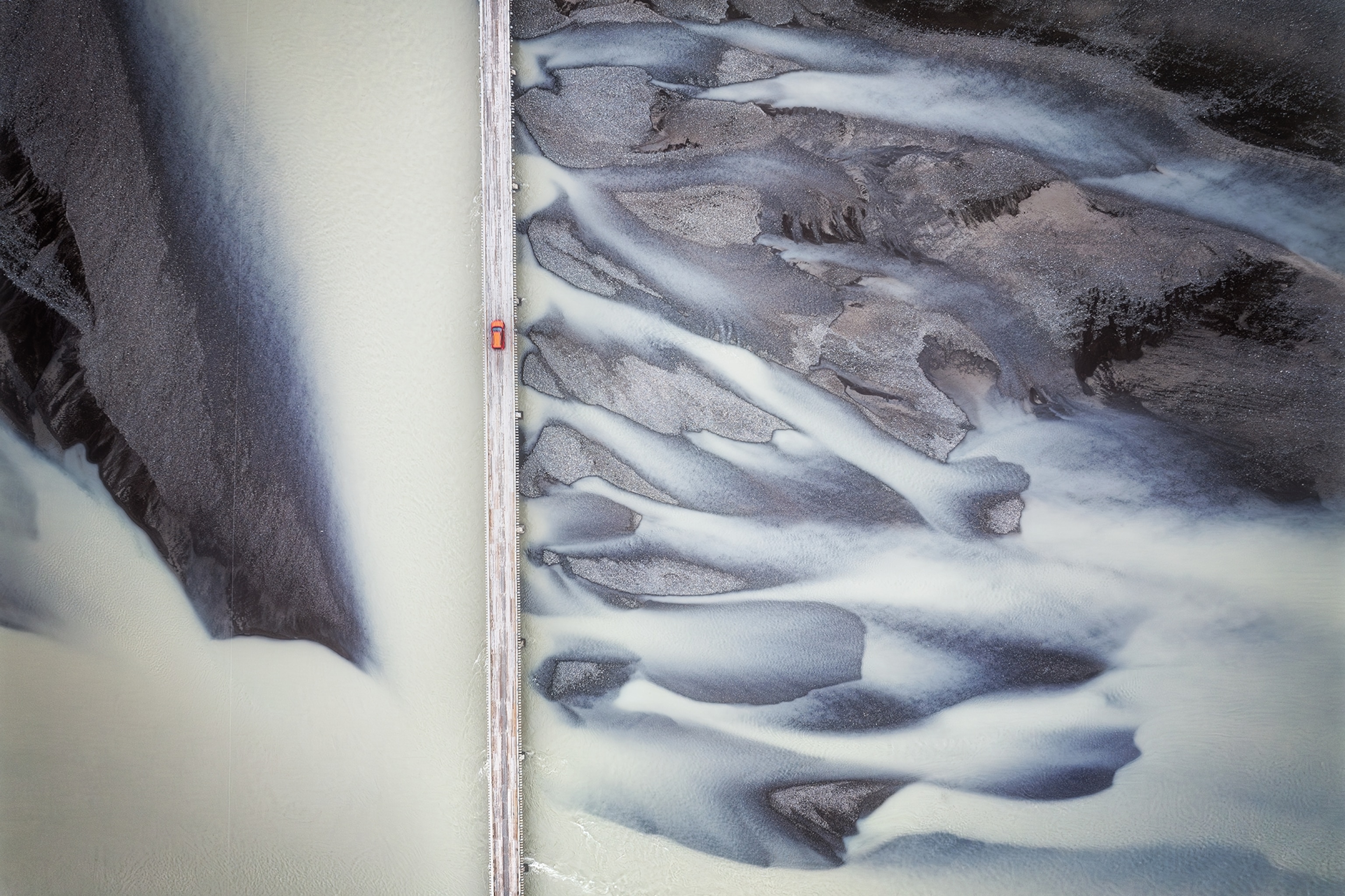 A glacial river in Iceland merging with the black sands of a local volcano as a car drives by on an empty road.