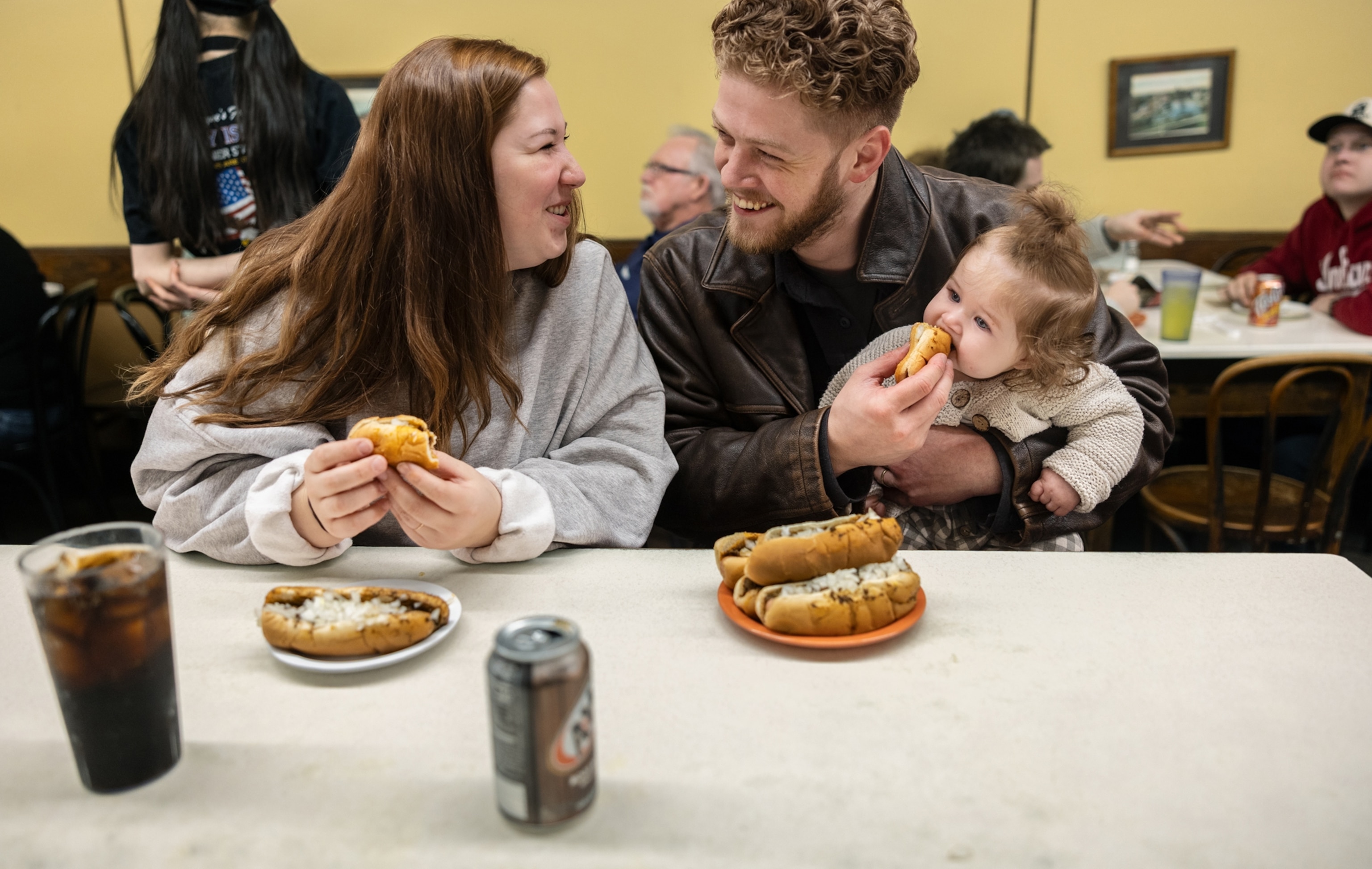 A mom on the left, and dad on the right holding their daughter, all eat hot dogs while sitting at a table in the restaurant