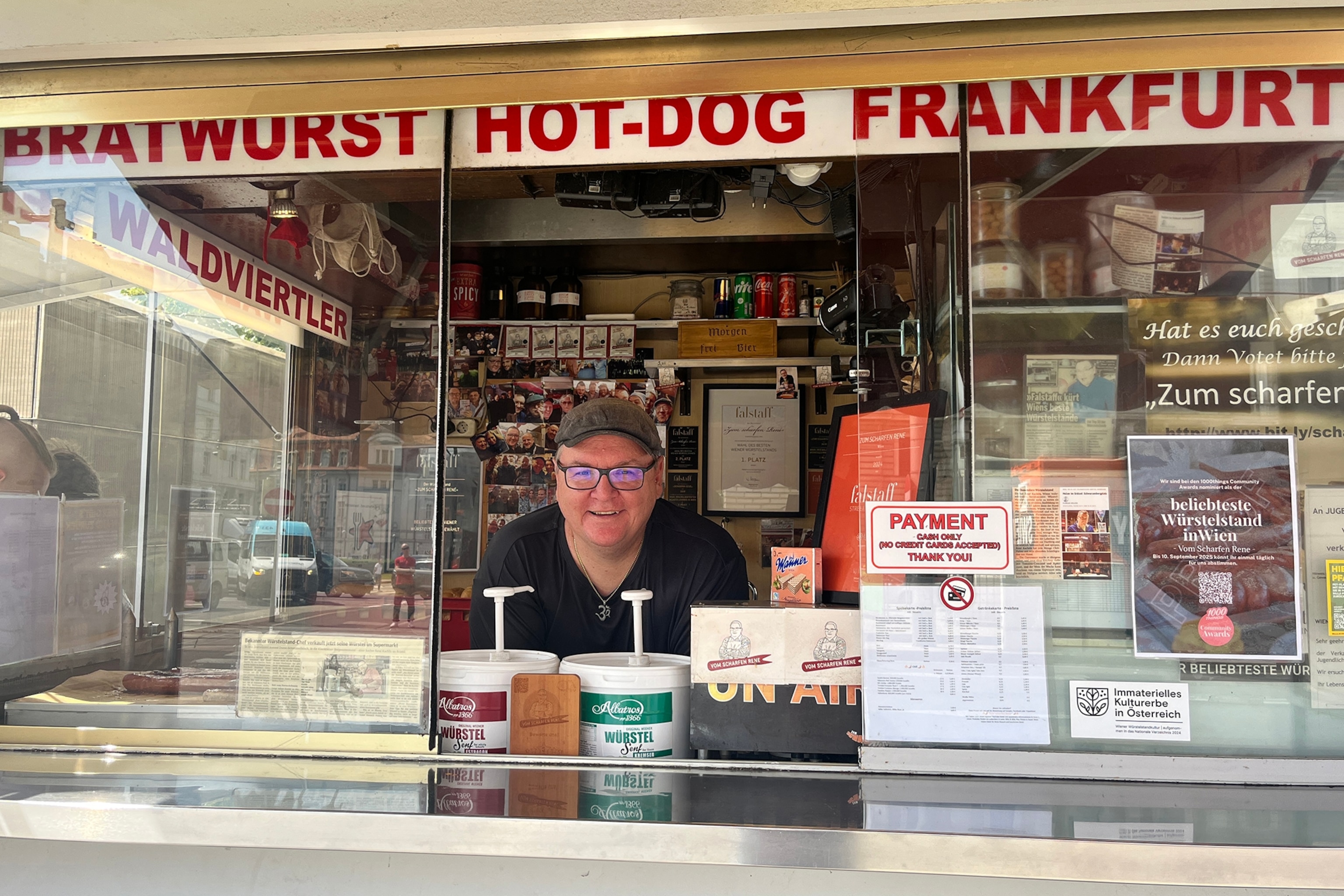 A smiling man leaning out of his food truck window, where he sells sausages with various condiments.