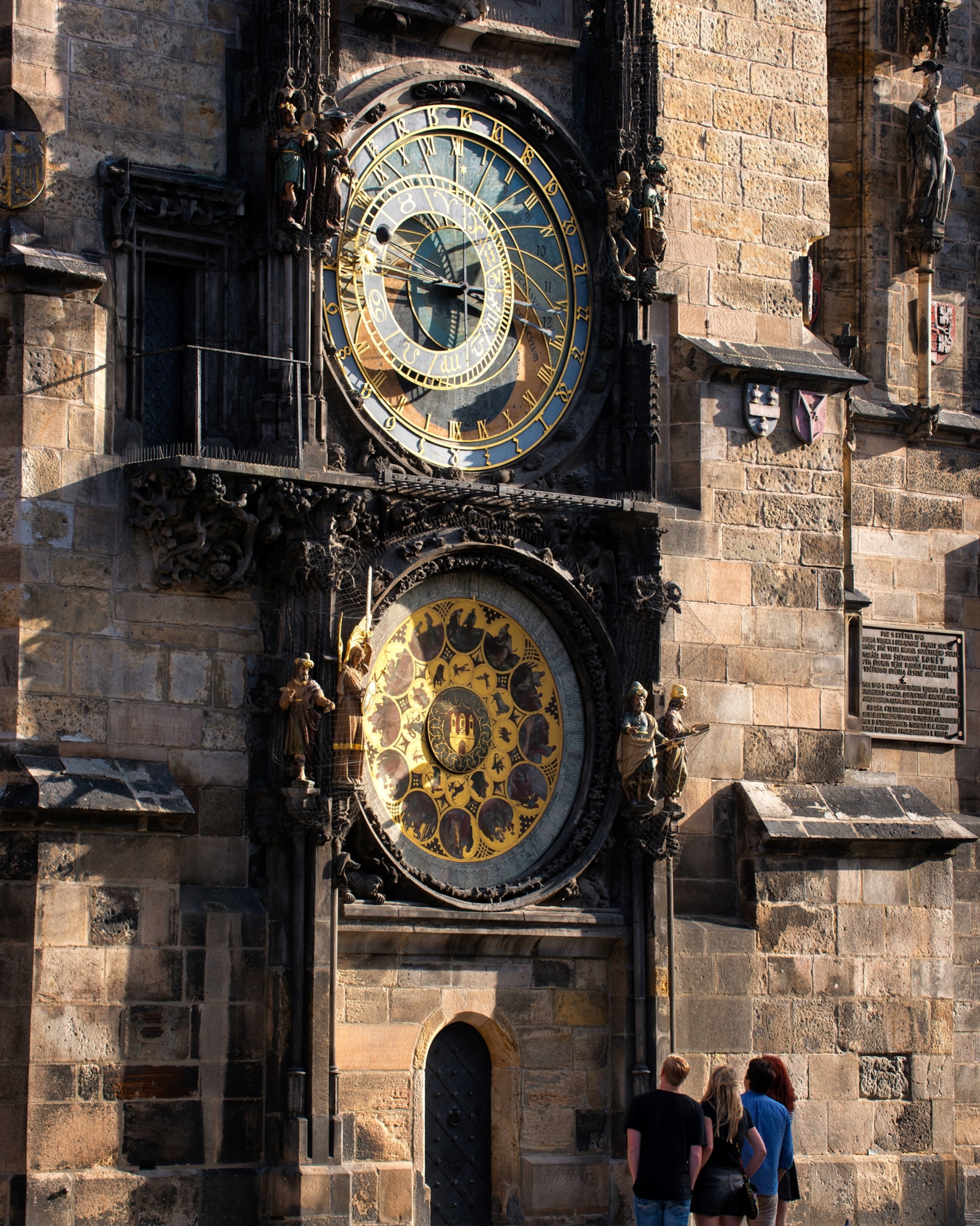 Historic astronomical clock on a sunlit stone wall, with intricate dials and zodiac symbols. Three people admire the details below.
