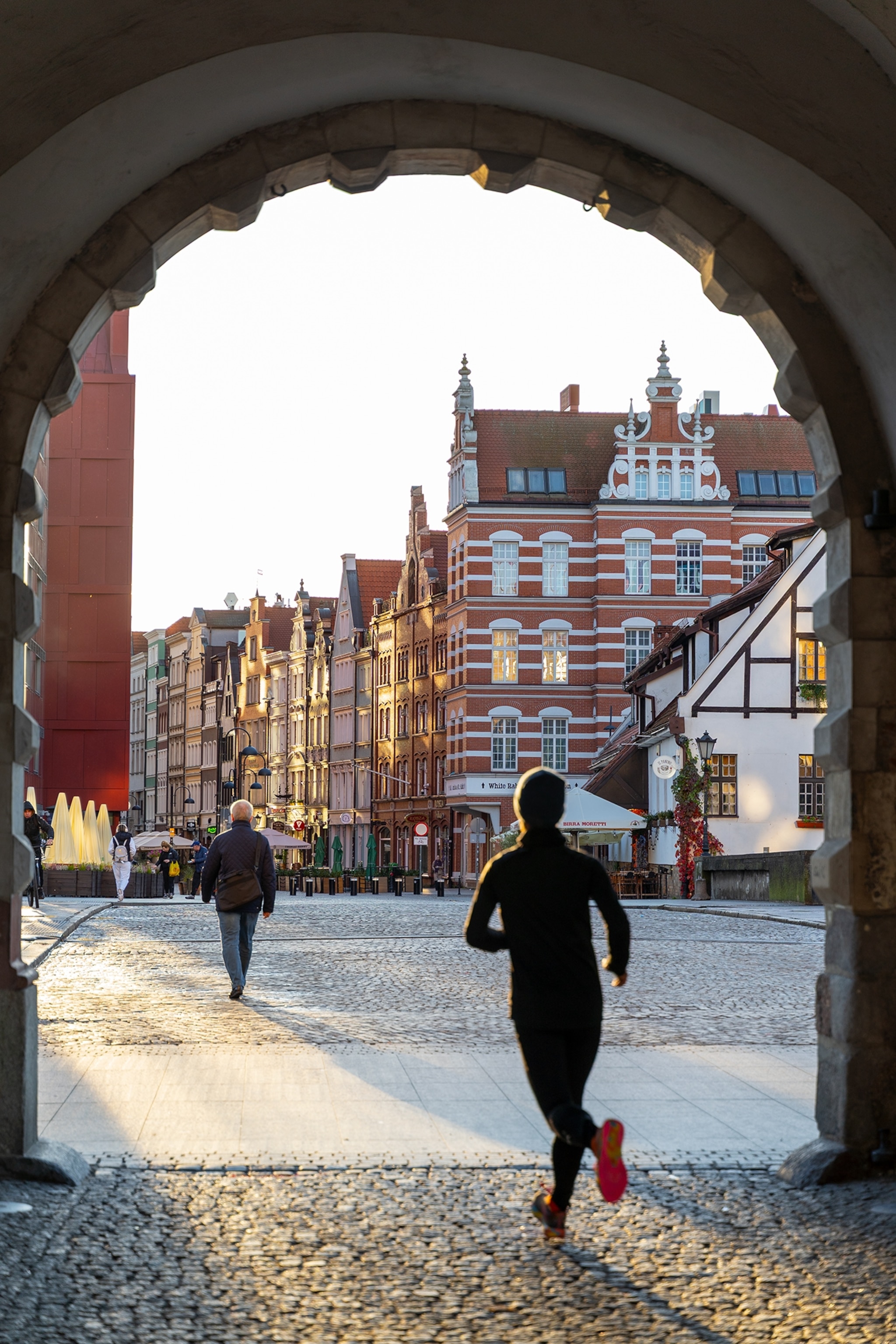 A person jogs through a stone archway into a historic city square lined with ornate buildings, cobblestone streets, and outdoor cafes in warm evening light.