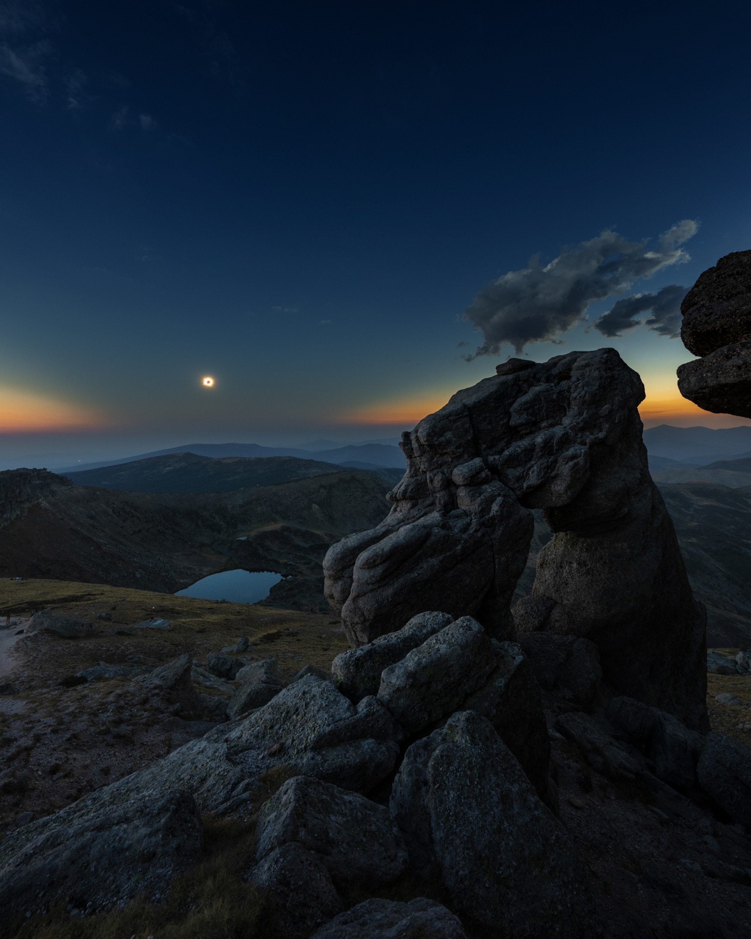 A mountain landscape beneath a dark sky during a total solar eclipse, with rugged foreground rocks, distant ridges, a small alpine lake, and wispy clouds