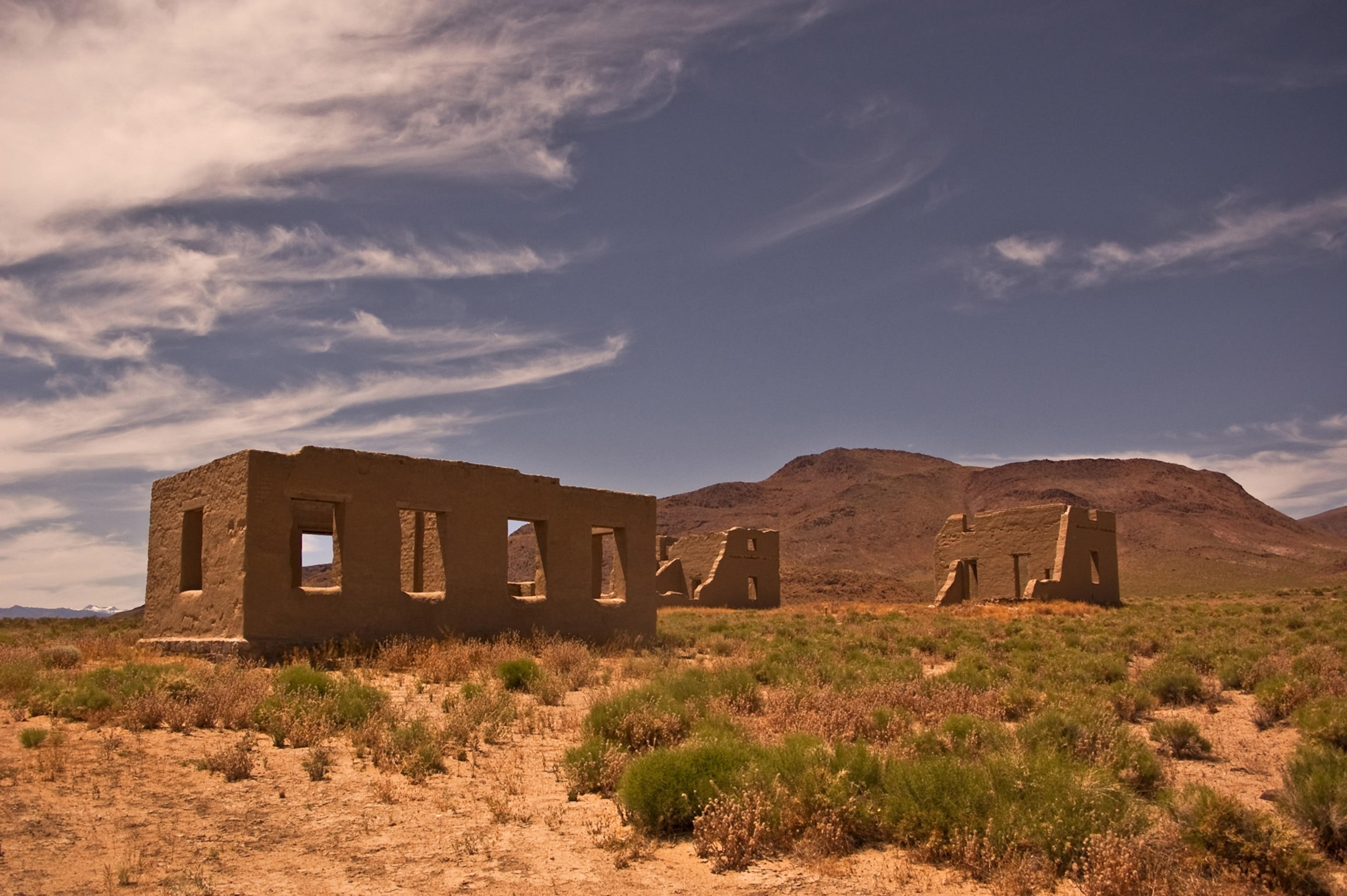 The empty ruins of a few small brown buildings stand alone in a desert landscape beneath a blue sky.