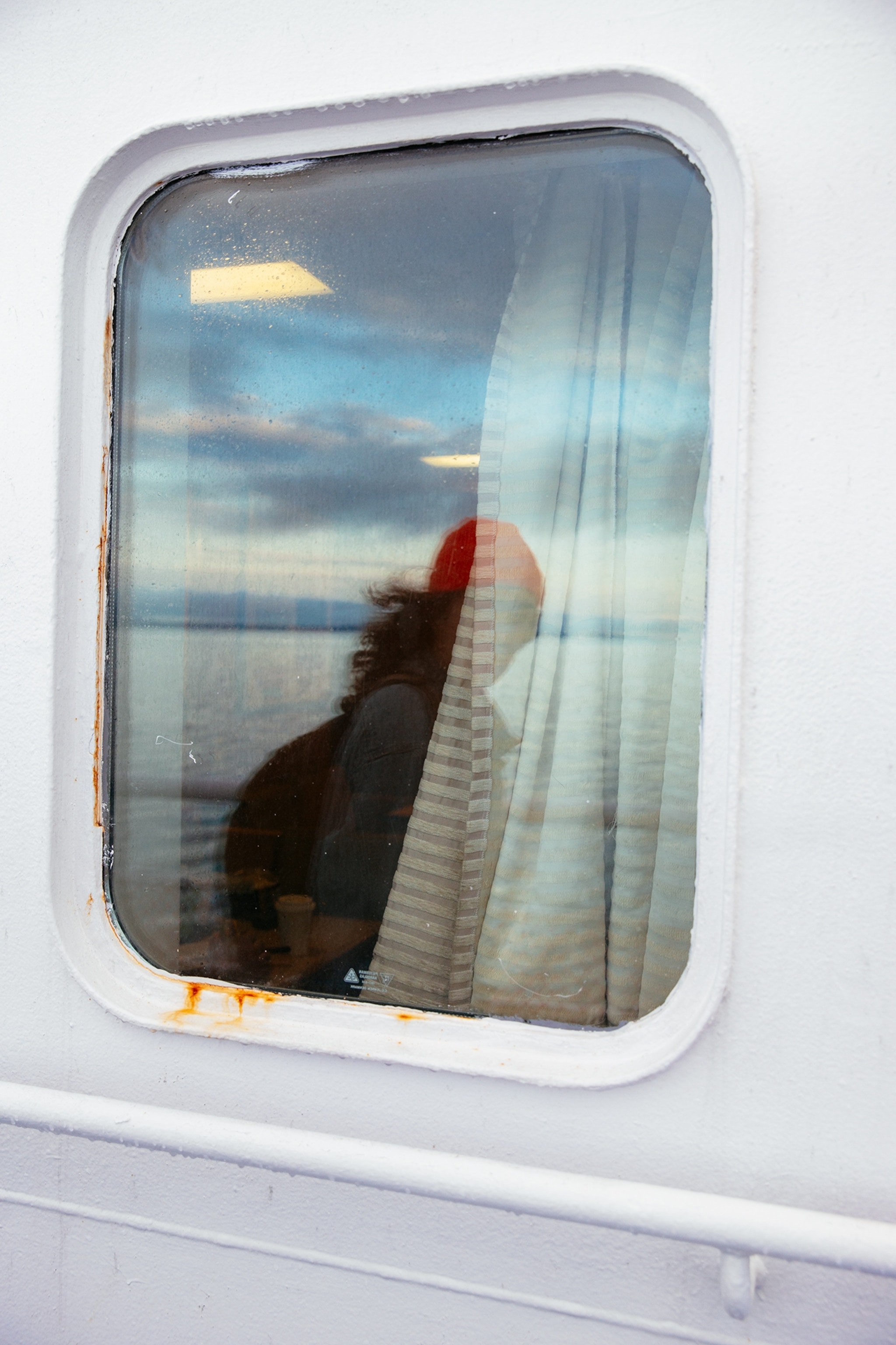 A person is seen through the window of a ship or ferry, with reflections of the water and sky visible on the glass.