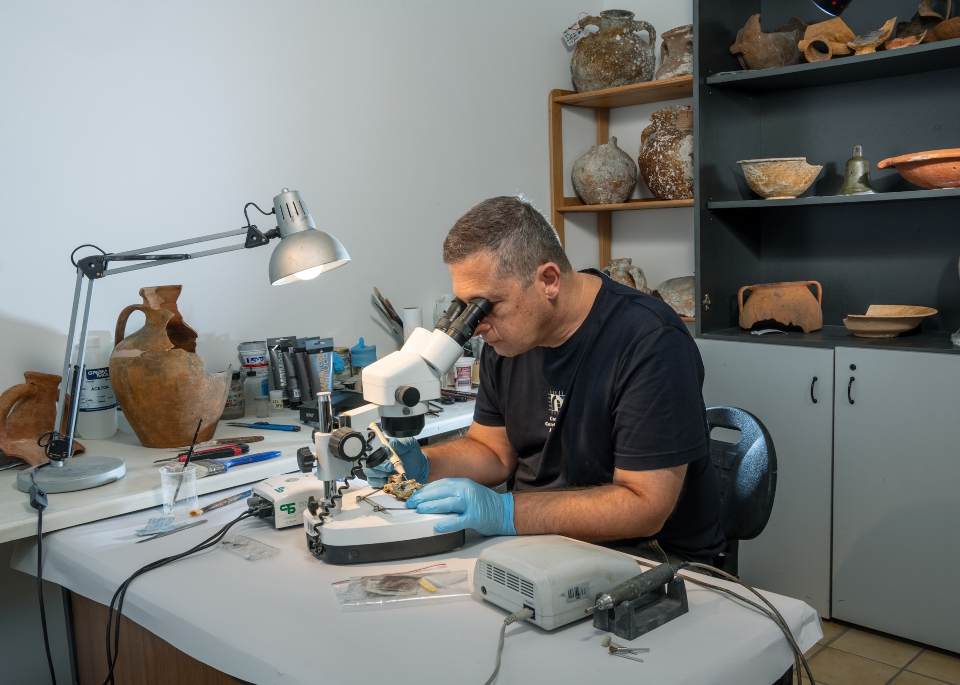 A person sits at a desk using a magnifier and lamp. Behind and next to the person are various artifacts and tools.