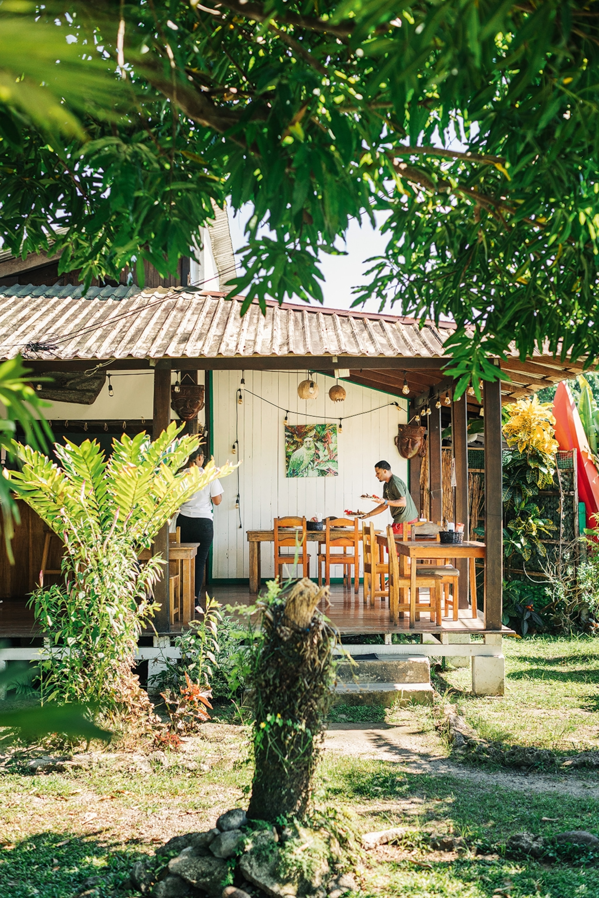 A terraced restaurant in a simple wooden bungalow with a vibrant garden and lanterns hung around the porch.