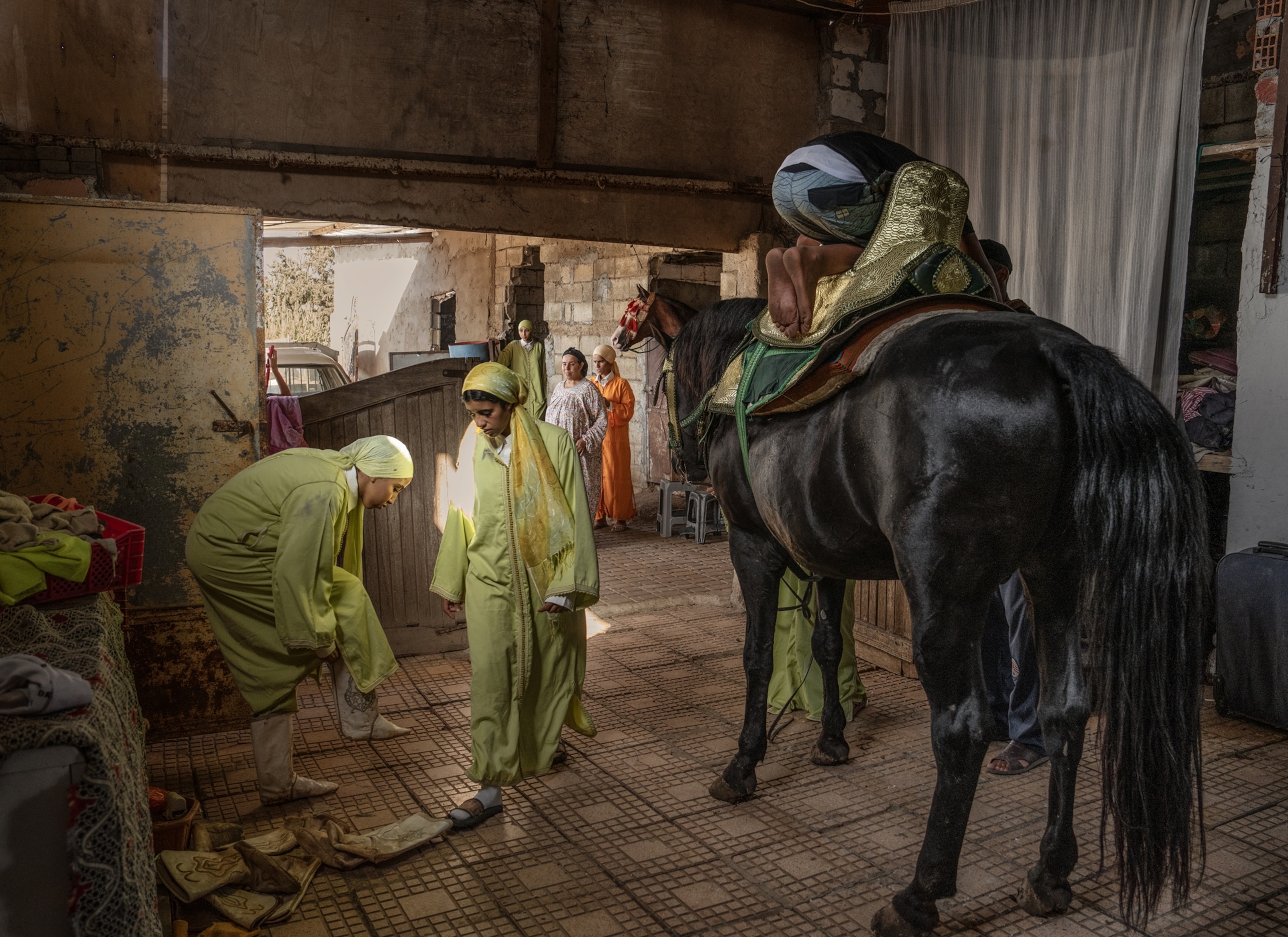Two performers get ready in green outfits. One is walking as the other pulls on their shoes. A horse stands next to them wearing a saddle and a person crouches on top of the saddle.