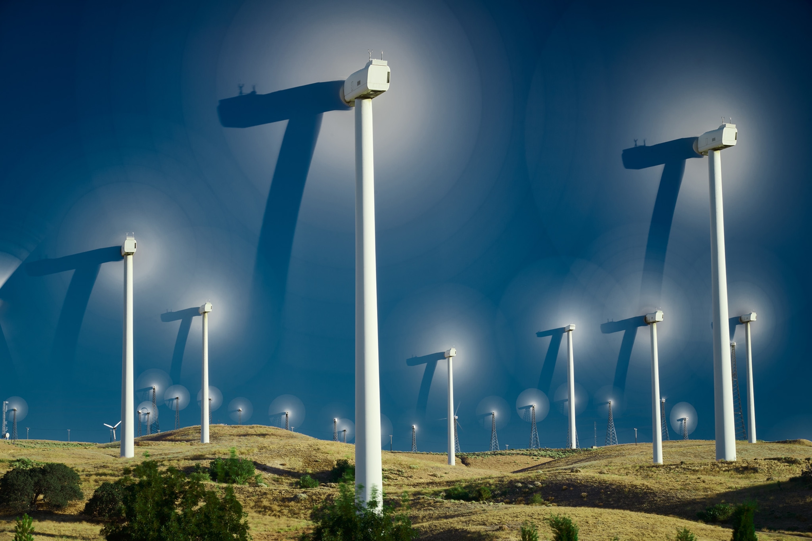 Tall white wind turbines spin fast, making their blades disappear against a dark blue sky. They stand among rolling green hills.
