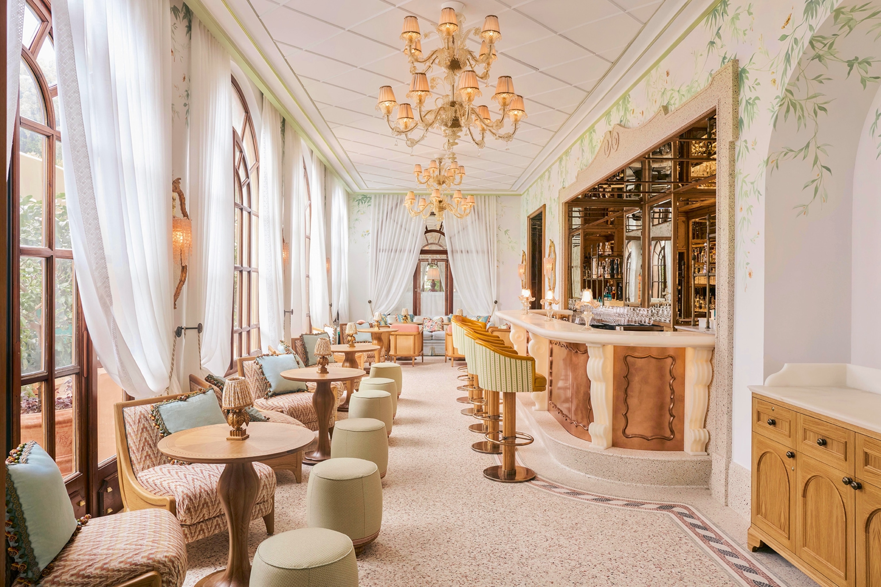 An airy, long bar facing large windows with draped curtains, arm chairs and chandeliers.