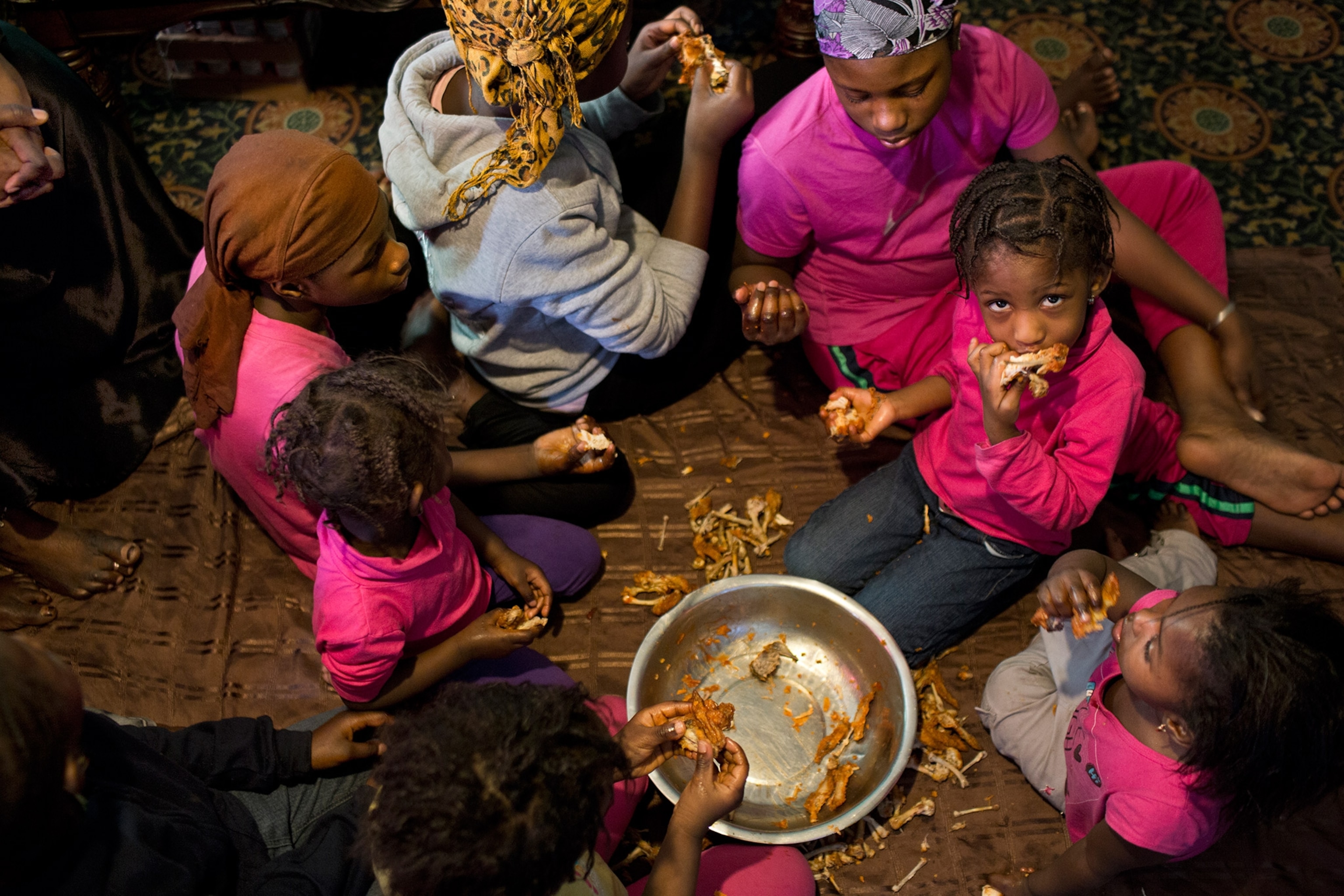 a group of children eating chicken out of a bowl on the floor of their family's apartment in the Bronx