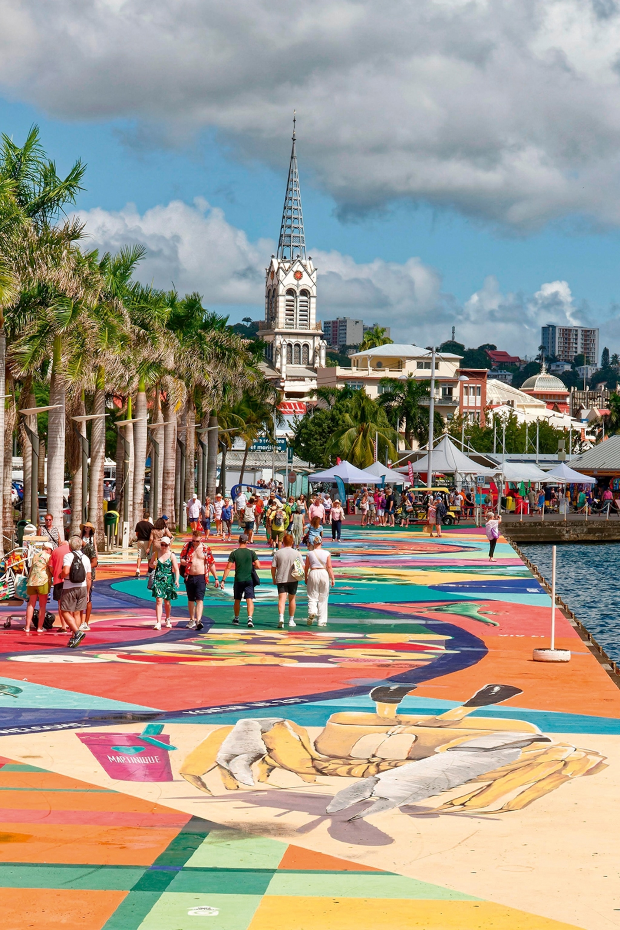 A busy promenade with a mural on the ground, palm trees along the city-side and a modern church steeple in the background.