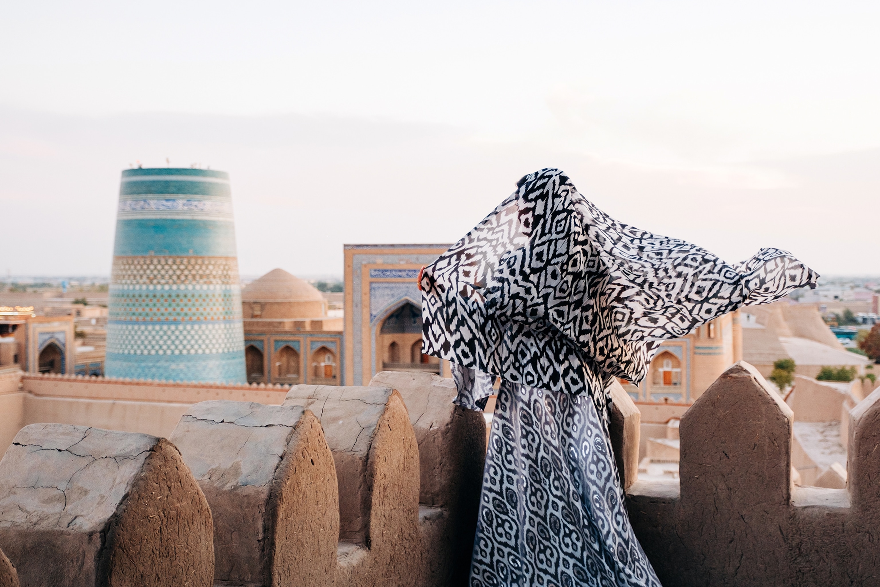 A woman with billowing tunic and head scarf from behind, looking over the landscape of a medieval city.