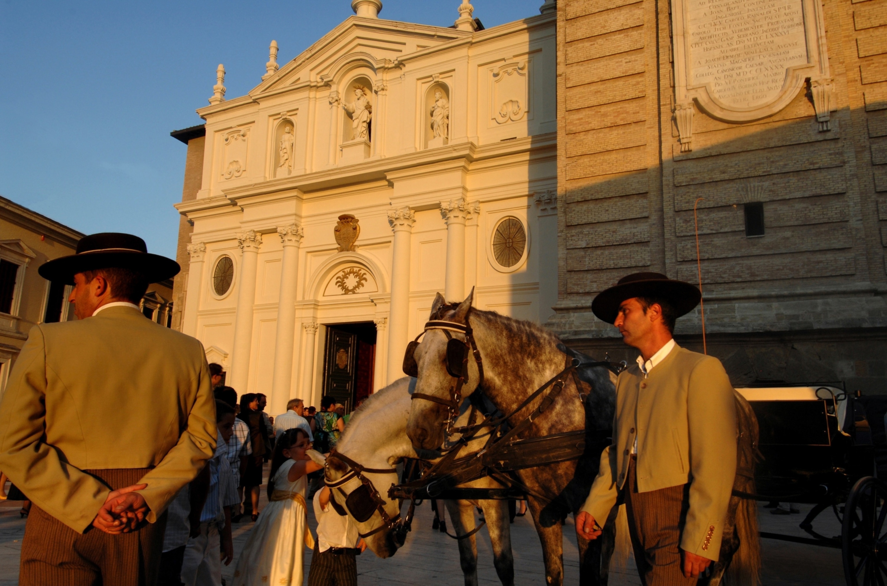 Men with wide brimmed hats holding a horse with a large brick building being highlighted by warm light in the background.