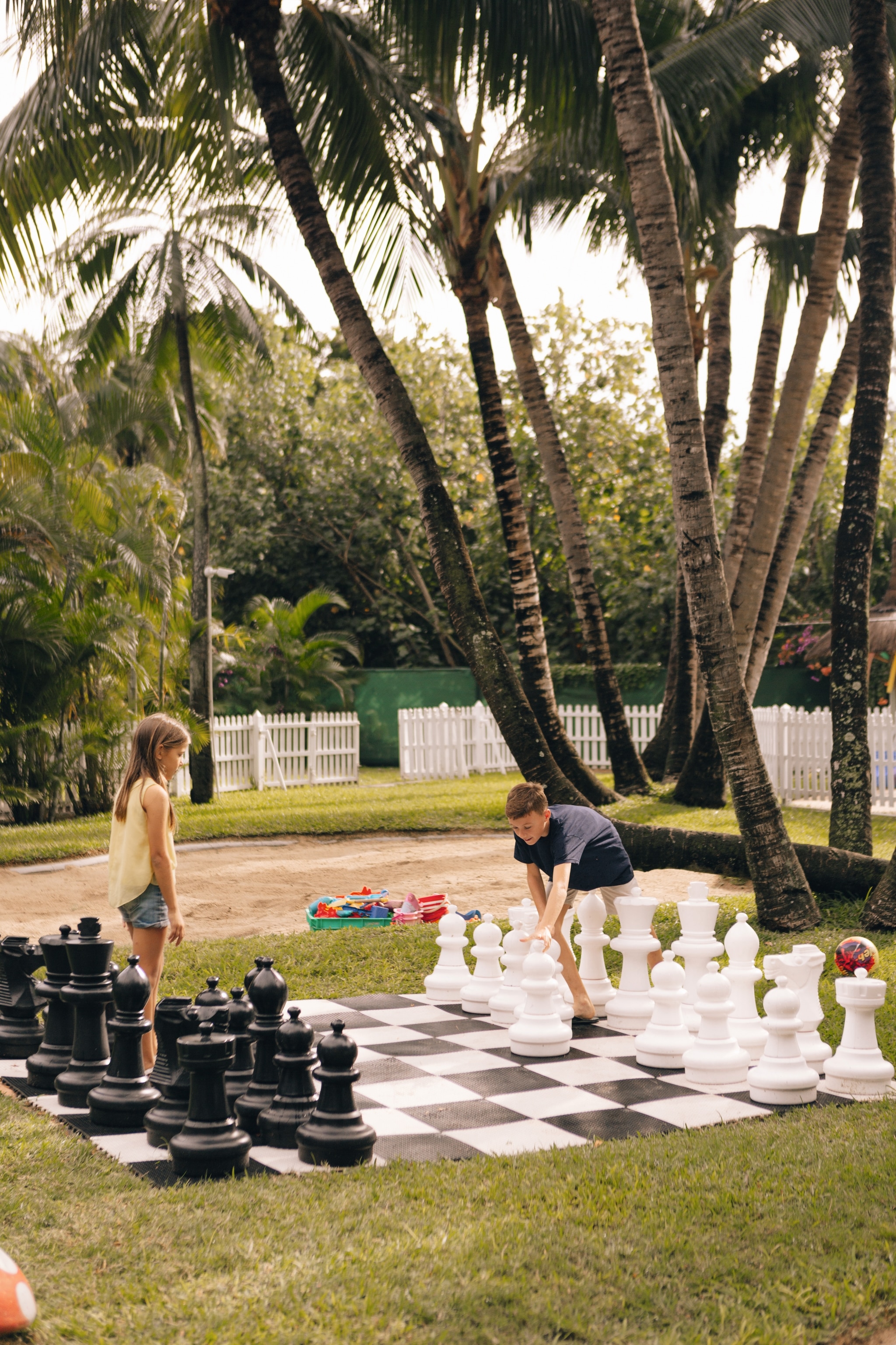 Two young children playing on a giant chessboard, in a tropical setting.